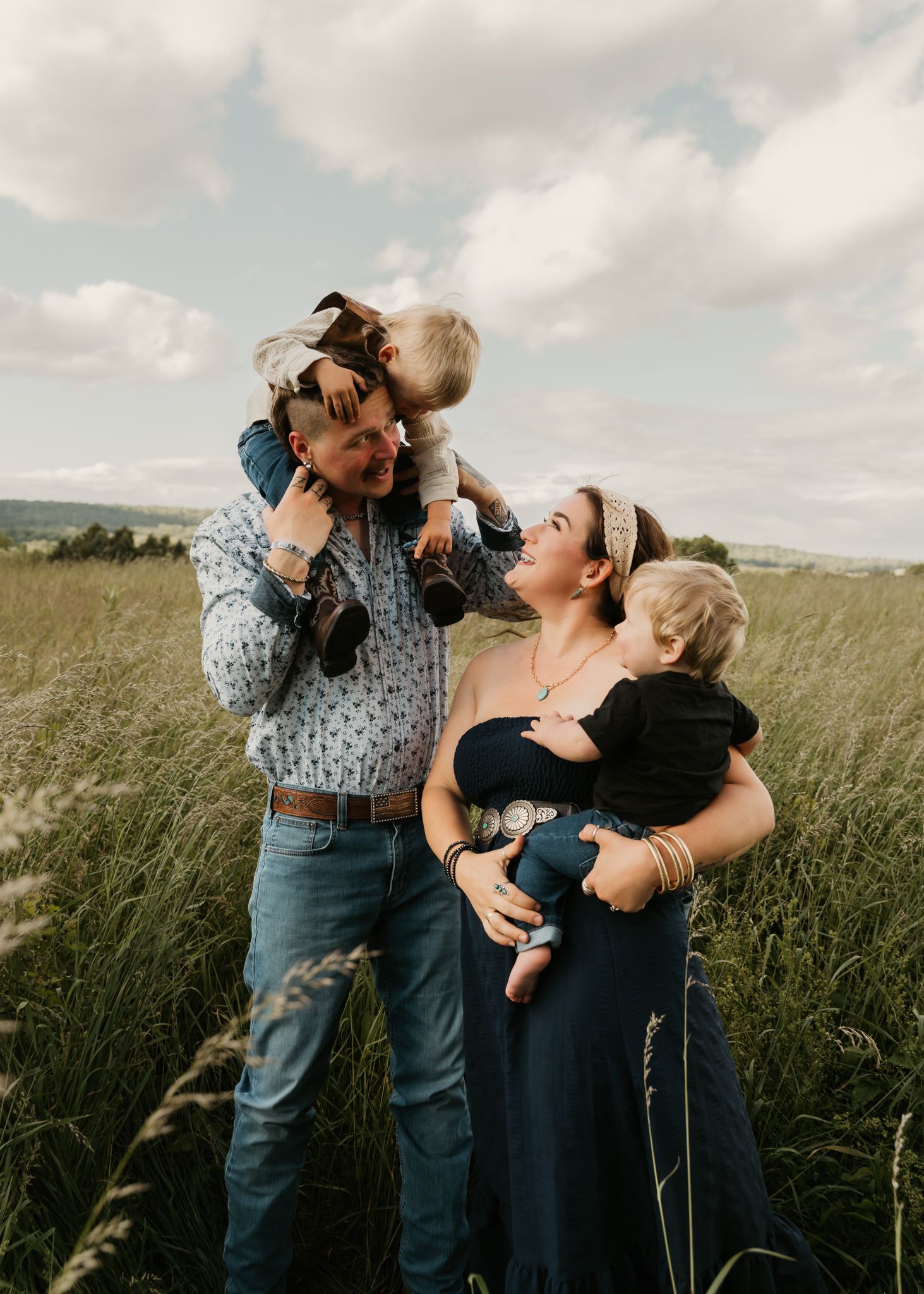 Joyful family of four in a tall grass field, father lifting child while mother holds baby.