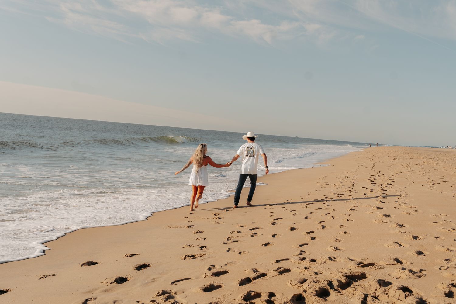 Couple walking hand in hand along a sandy ocean beach with waves and clear sky.