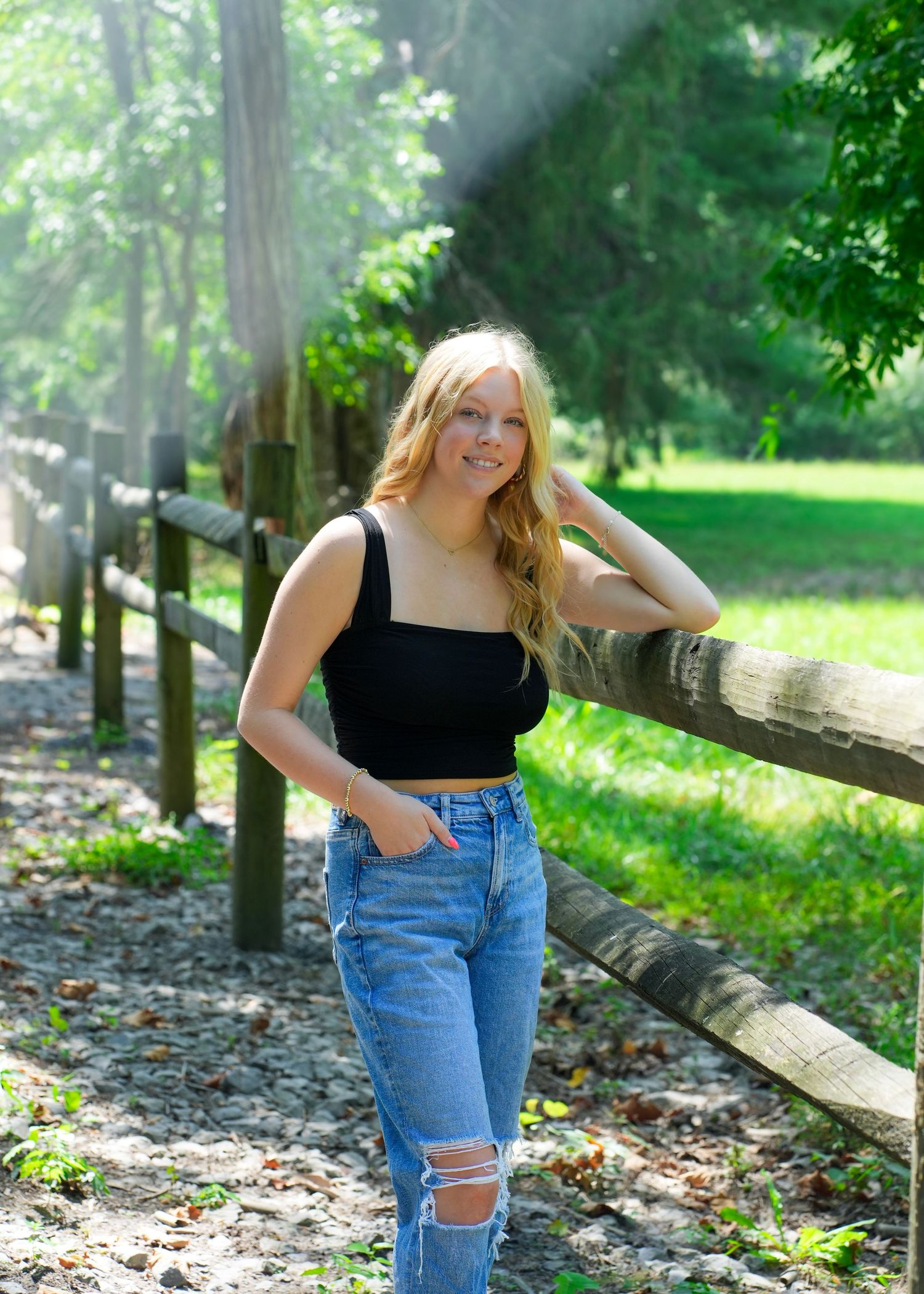 Blonde teenage girl in black top and jeans leans against wooden fence in sunlit forest.