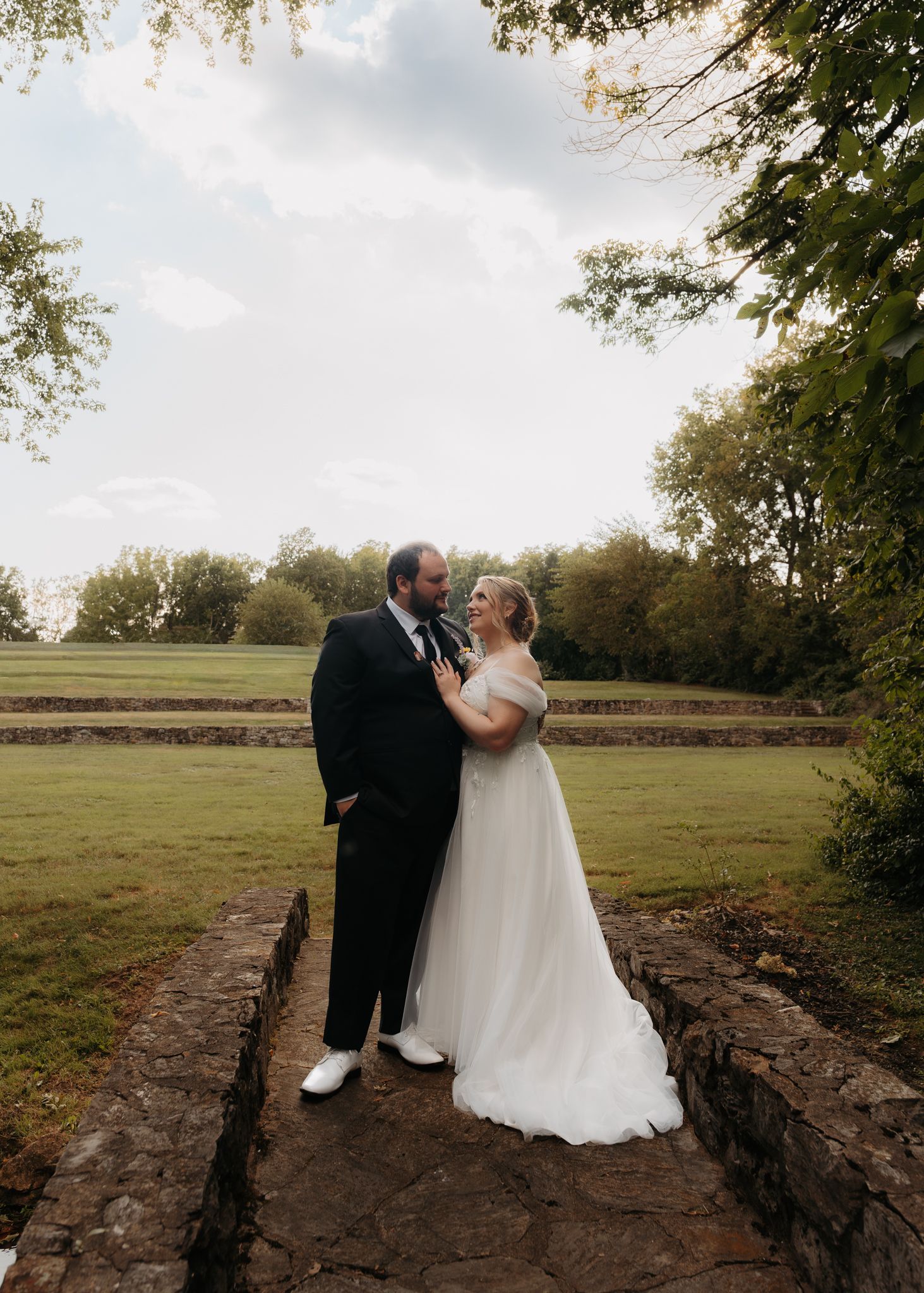Bride and groom share romantic moment on stone terrace surrounded by lush greenery under dramatic cloudy sky.