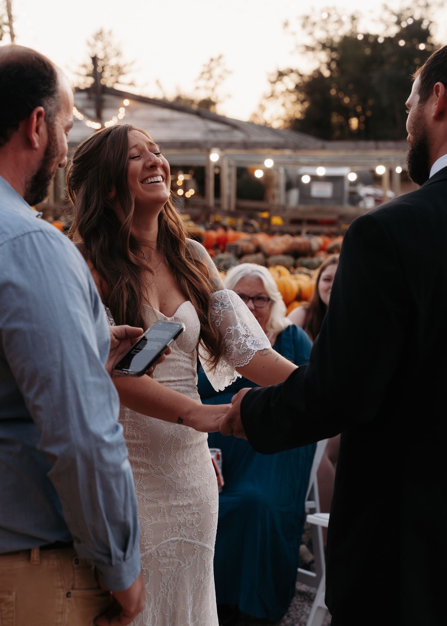 Joyful bride laughing while holding hands with groom during outdoor wedding ceremony at dusk.