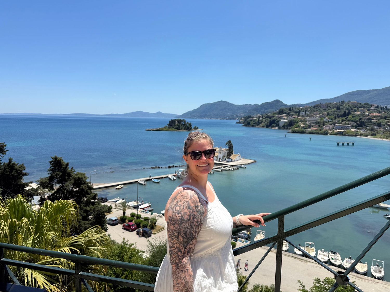 Woman with tattoos posing on a balcony overlooking a stunning blue Mediterranean harbor.