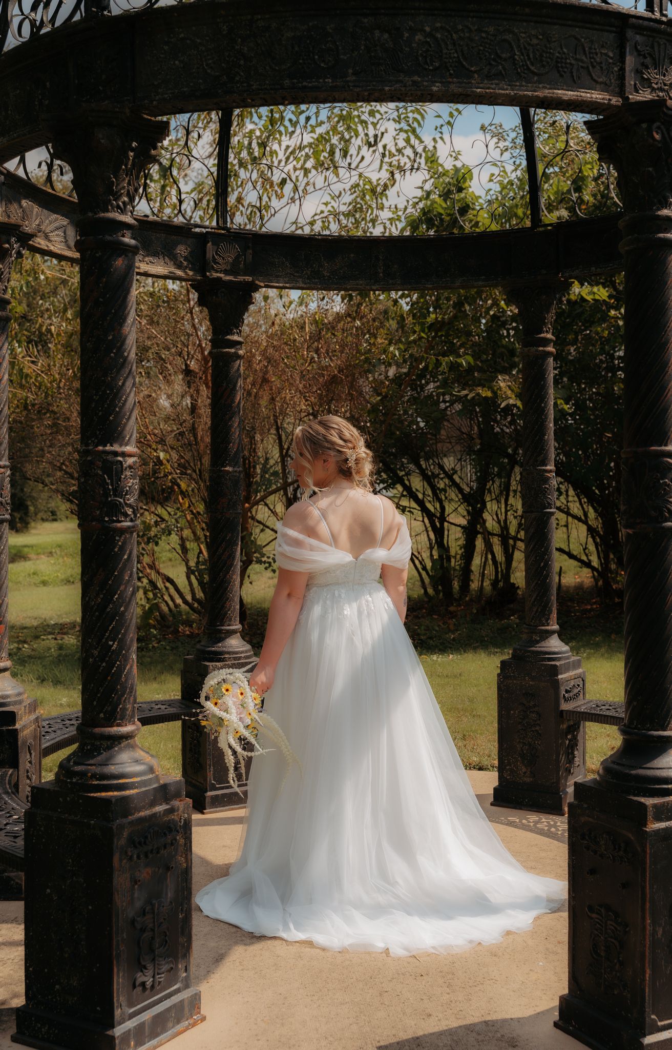 Bride in off-shoulder white tulle gown stands under ornate iron gazebo in garden venue holding bouquet.