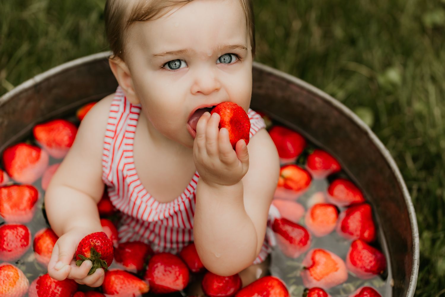 Adorable blue-eyed baby sits in a tub filled with fresh strawberries outdoors, happily eating a strawberry.