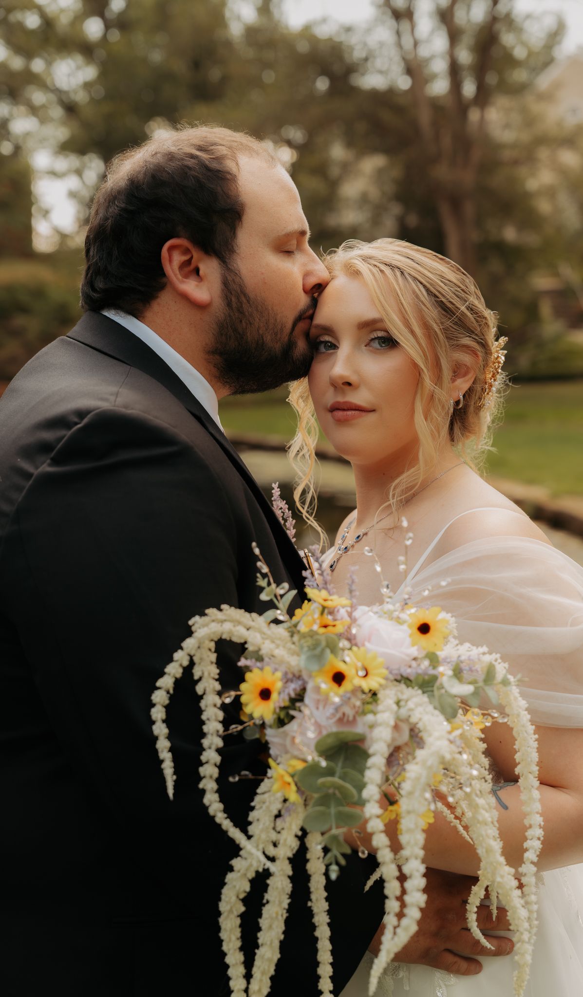 Groom in dark suit kissing bride on temple as she holds cascading sunflower and pearl wedding bouquet.