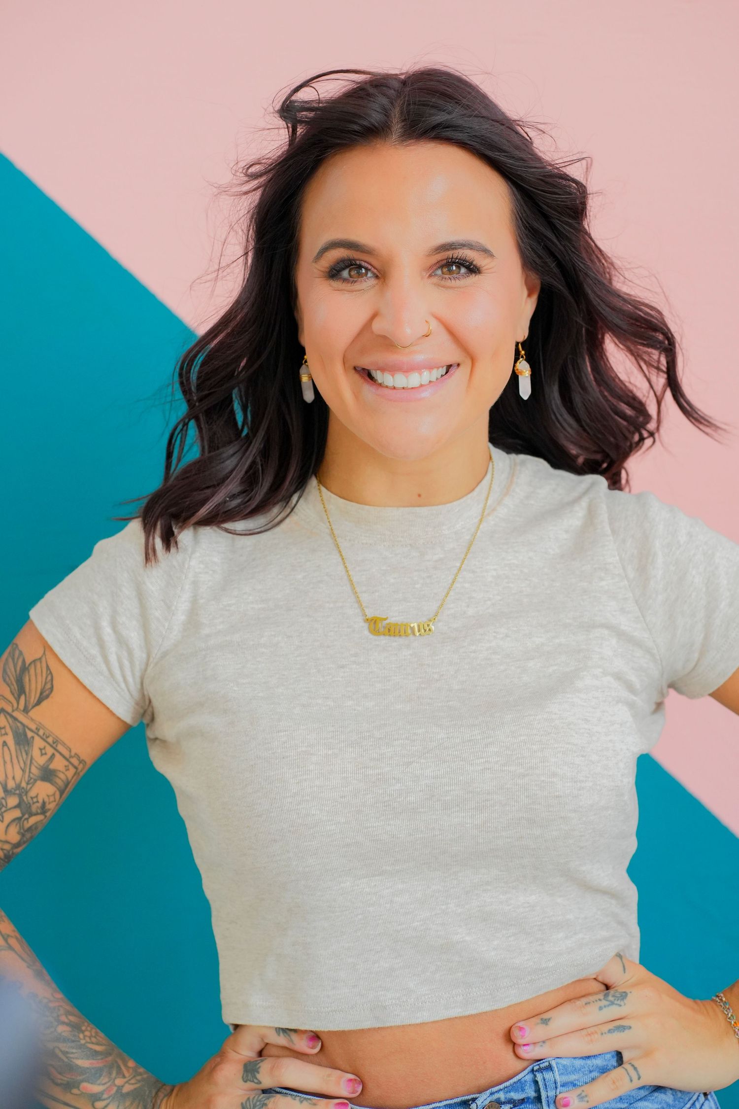 Smiling brunette woman with tattoos in grey crop top poses against colorful pink and teal wall.