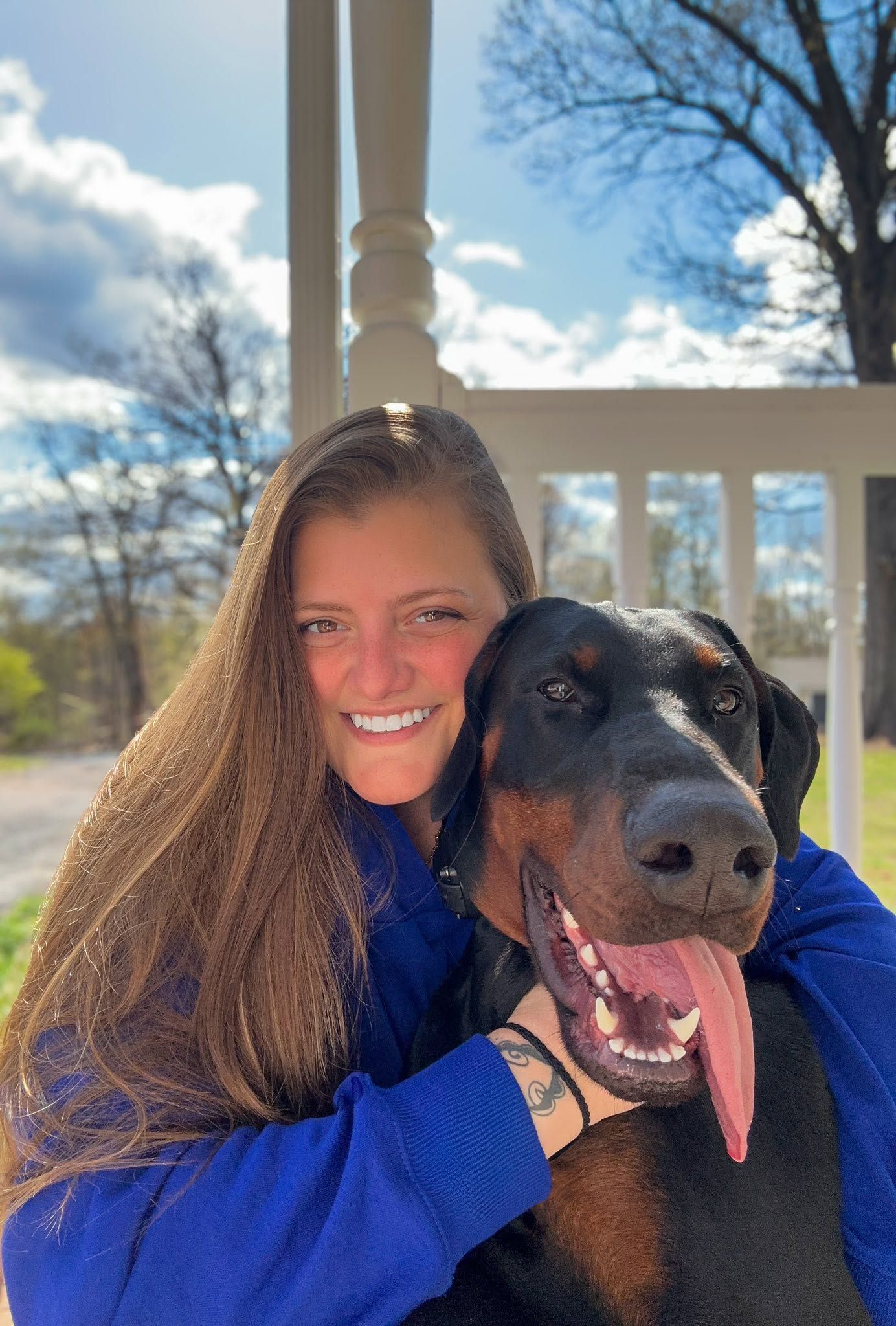 A smiling young woman with long brown hair hugs a happy Doberman Pinscher on a sunny porch.