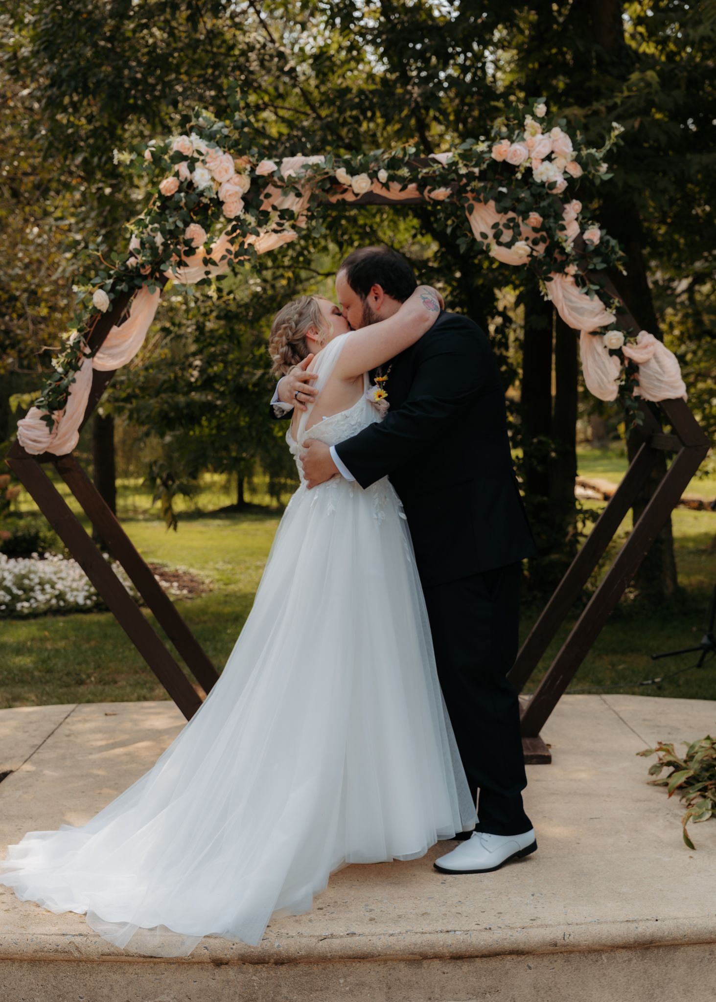 Bride and groom sharing first kiss under a floral hexagonal wooden arch in a garden.