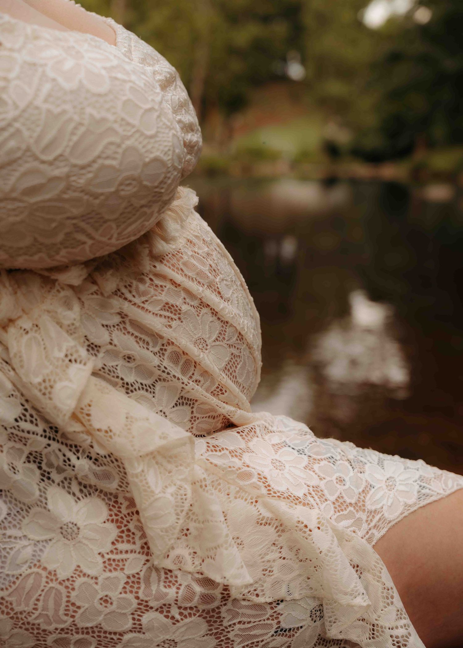 Close-up of a pregnant woman in a lace dress outdoors near a pond.