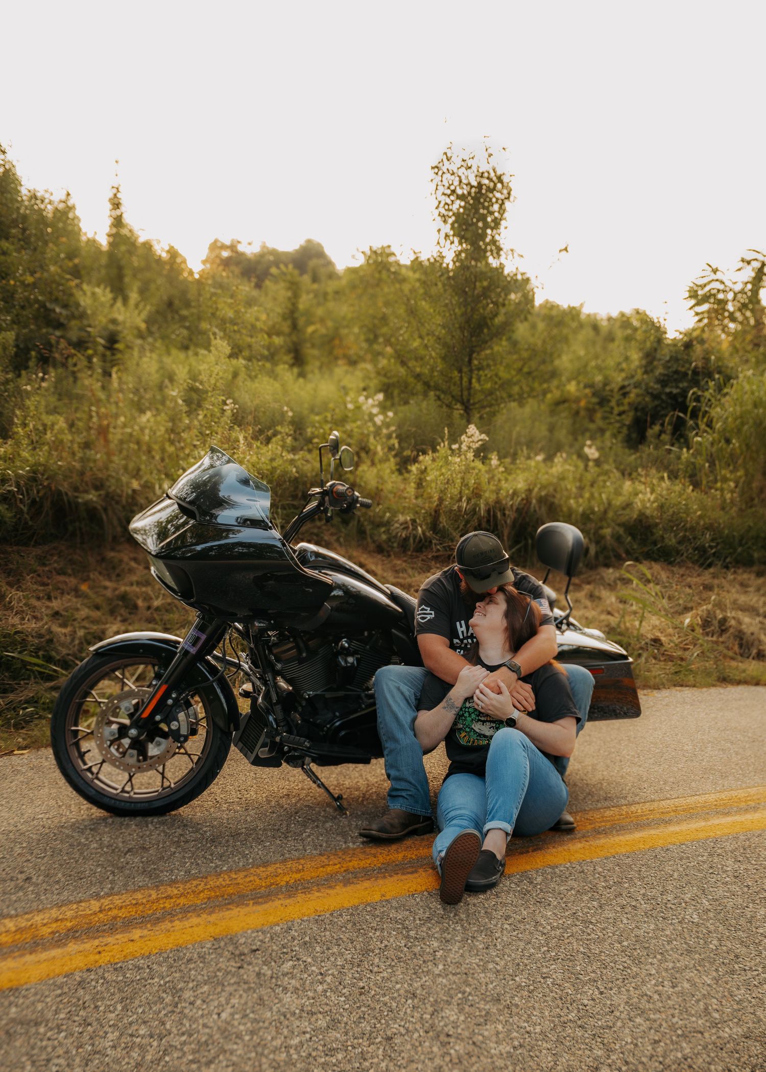 Couple sitting on road beside black Harley-Davidson motorcycle, sharing romantic moment with lush green trees in background.