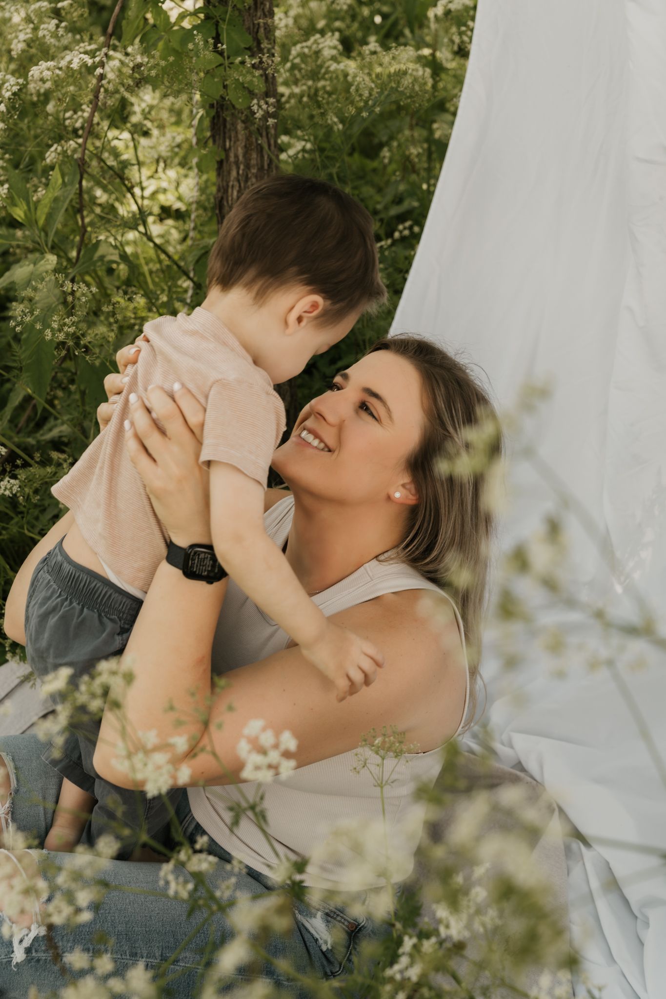Smiling mother holding young toddler boy near white fabric backdrop surrounded by green foliage.