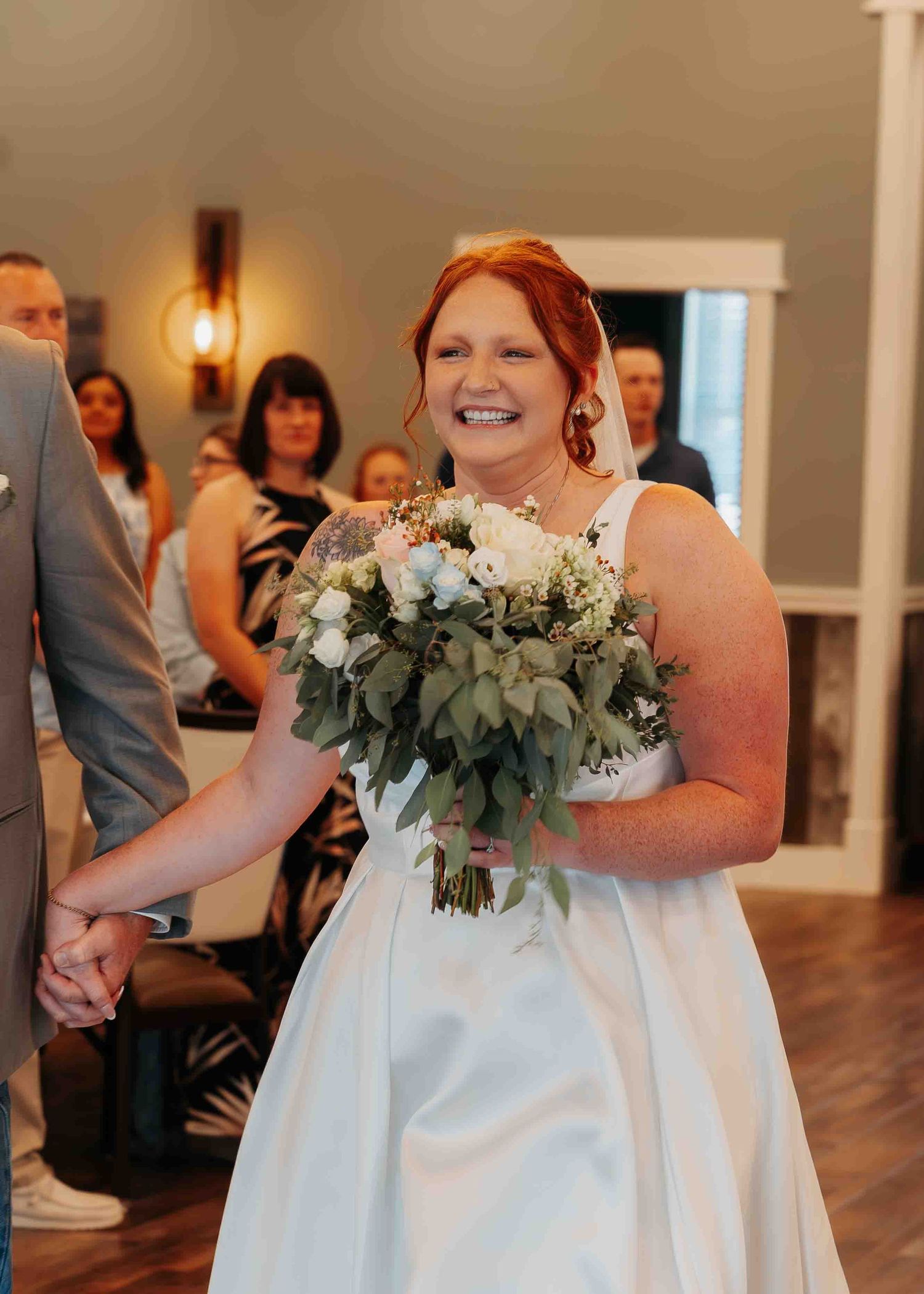 Smiling red-haired bride in white gown holds white and green bouquet while walking down the aisle at wedding.