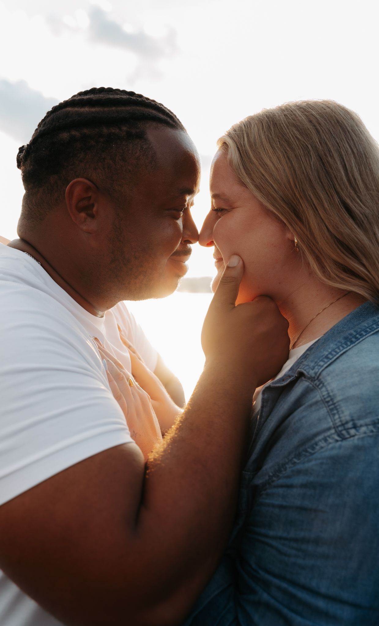 Interracial couple sharing an intimate nose-to-nose moment backlit by golden sunset light near water.