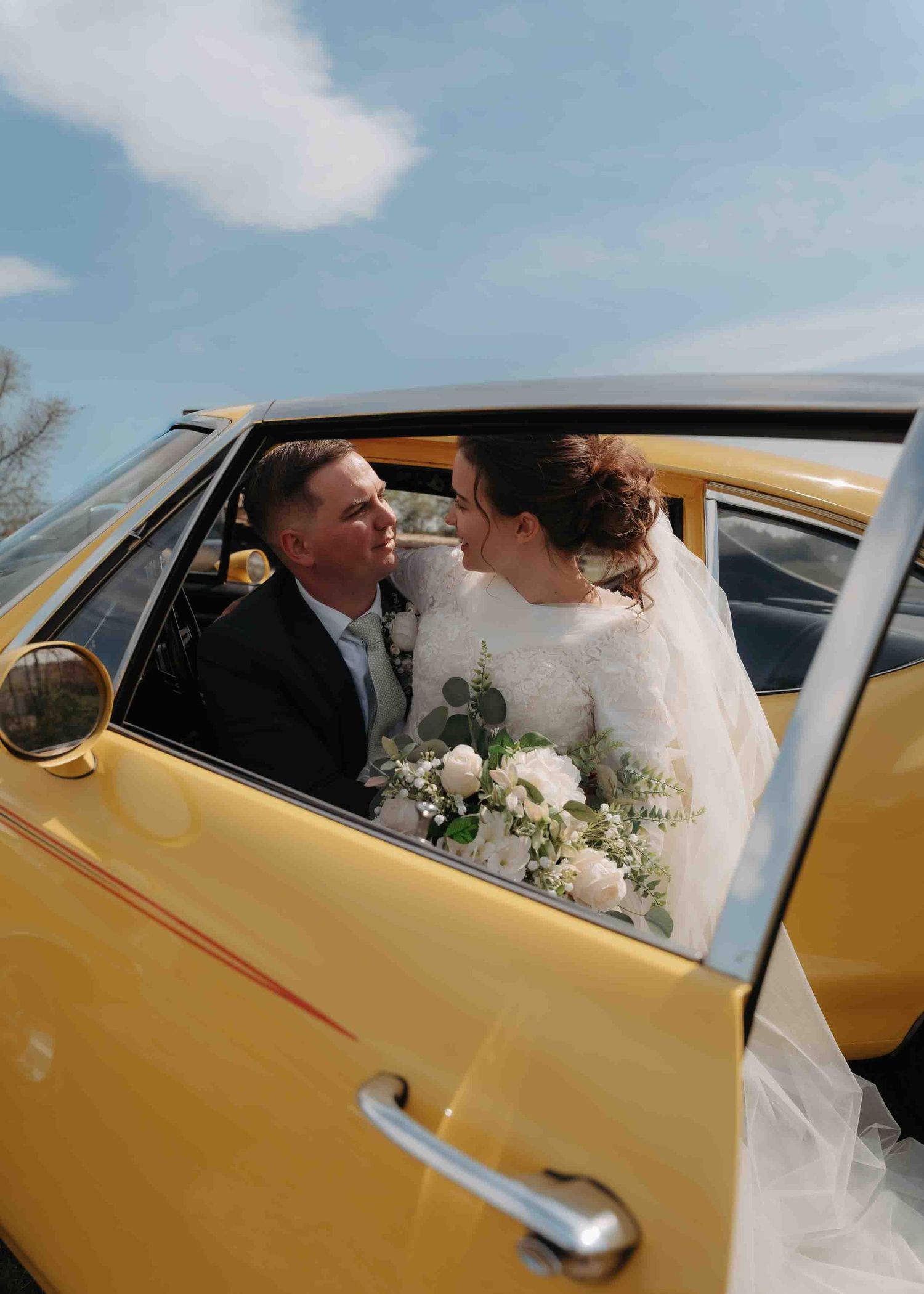 Bride and groom share a romantic moment inside a vintage yellow car on their wedding day.