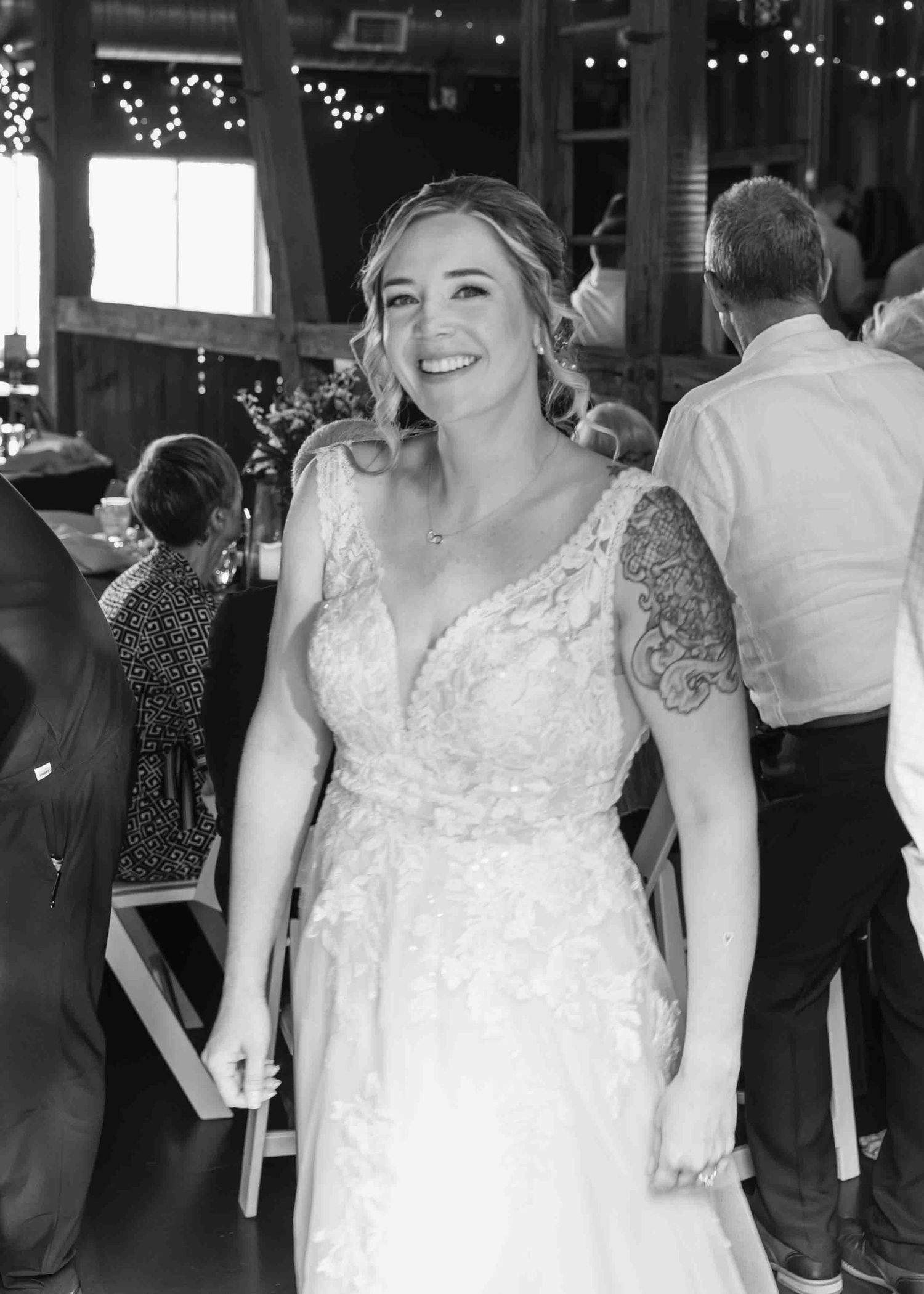 Smiling bride in lace wedding gown with arm tattoo at rustic barn reception venue.