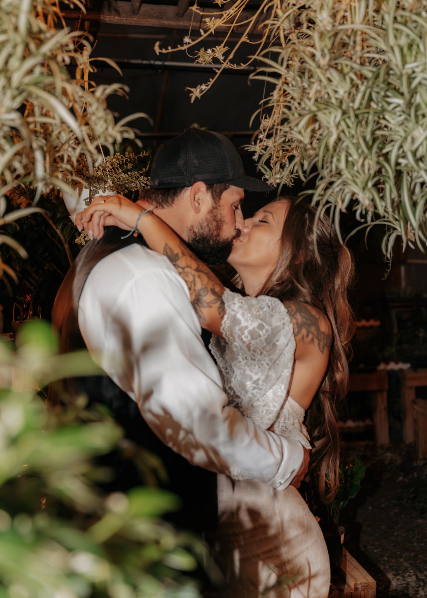 A couple shares a romantic kiss surrounded by lush greenery, with the groom in a white shirt and black cap embracing his tattooed bride.