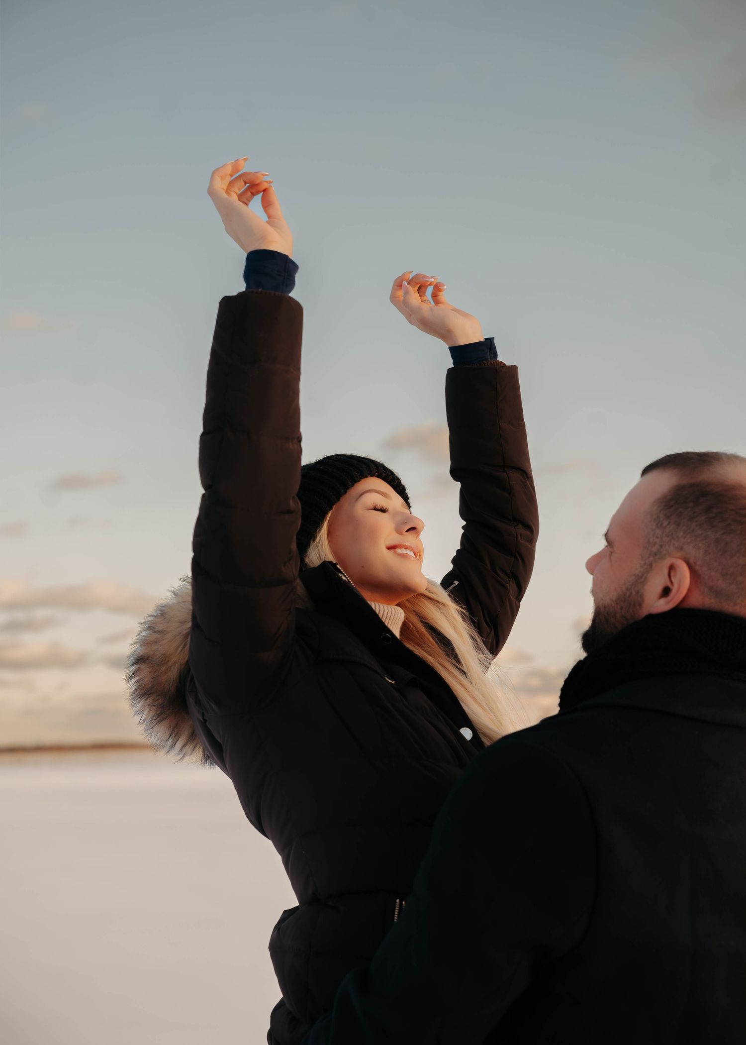 Couple celebrating outdoors in winter, woman raising arms joyfully against a snowy sky.