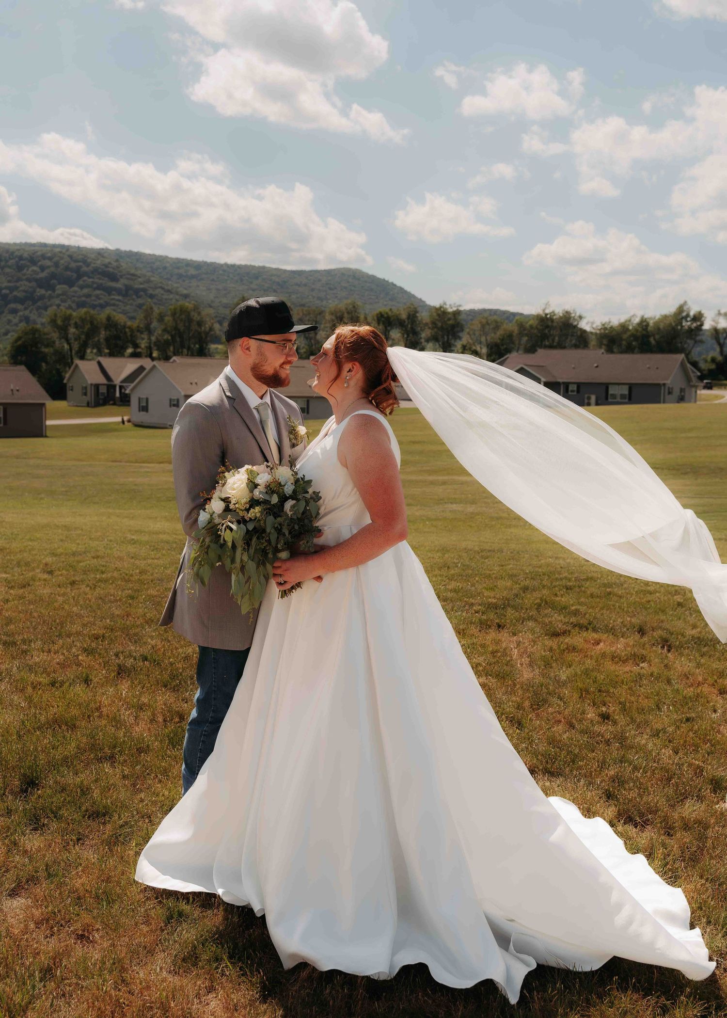 Bride and groom share romantic moment in open field with veil blowing in wind and mountain backdrop.