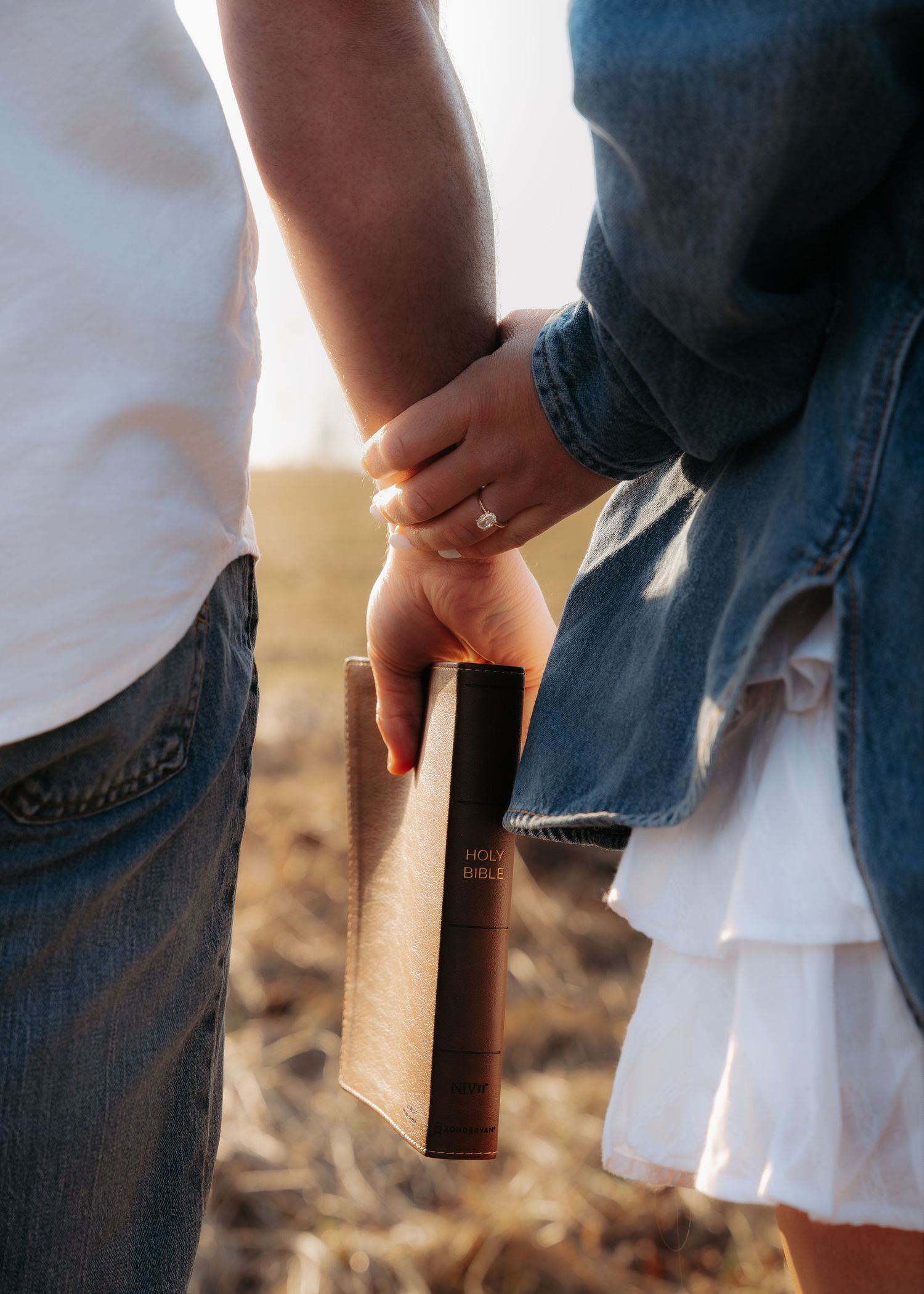 Couple holding hands and a Bible together in a field, woman wearing an engagement ring.