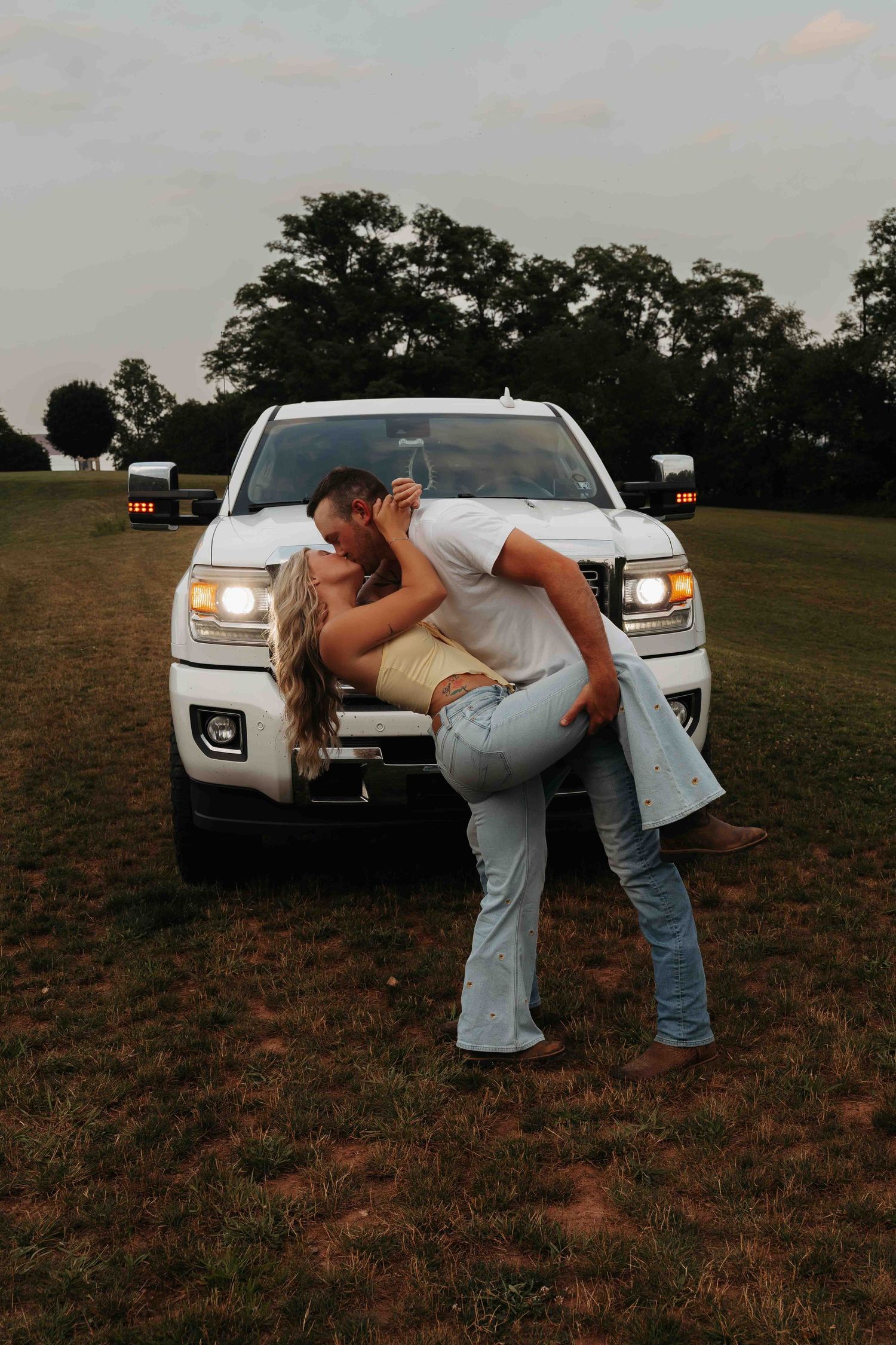 Couple kissing in a dip pose in front of a white pickup truck at dusk.
