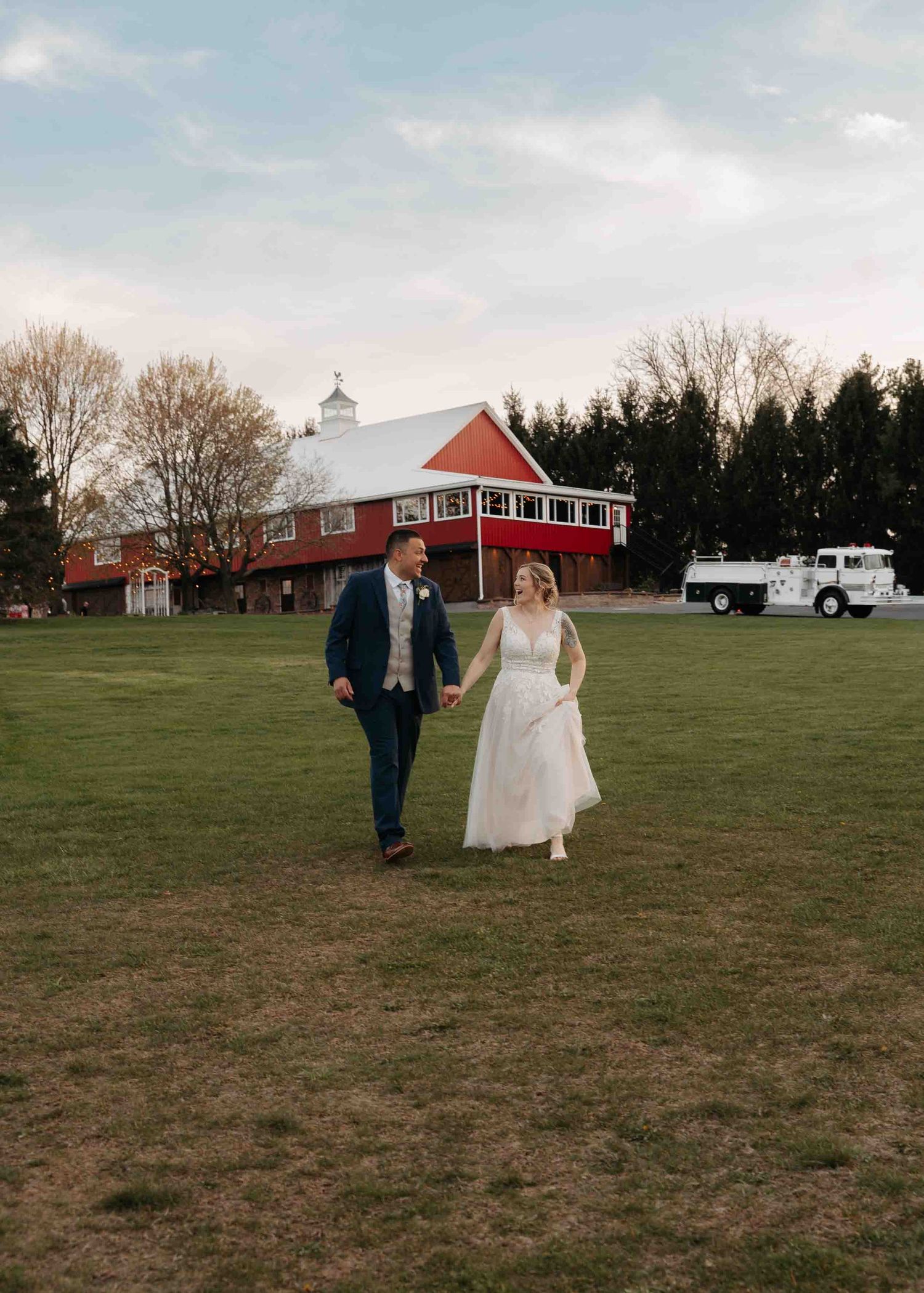Bride and groom walking hand in hand in front of red barn wedding venue.