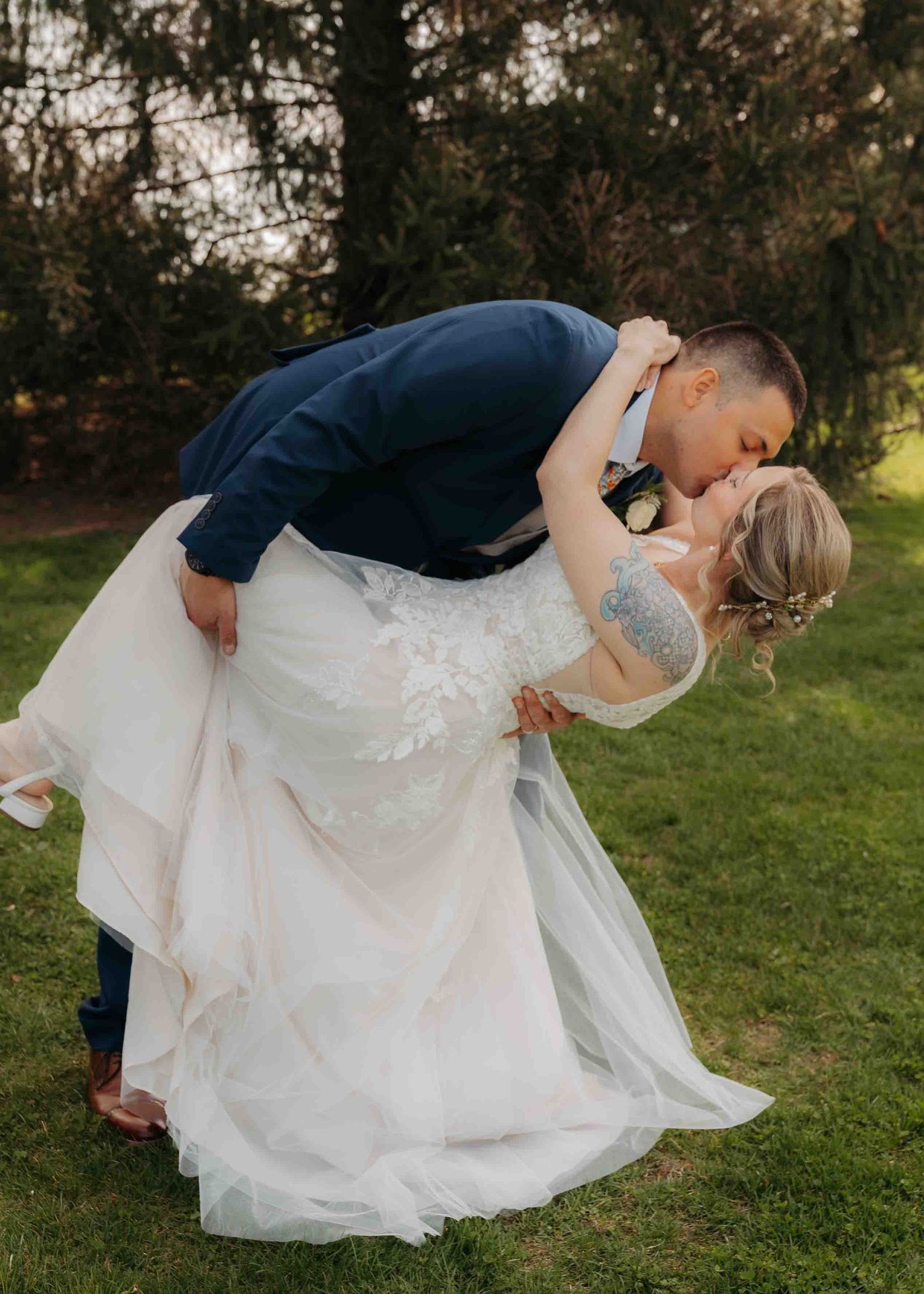 Groom dips bride for a romantic kiss on green lawn surrounded by lush trees at outdoor wedding.