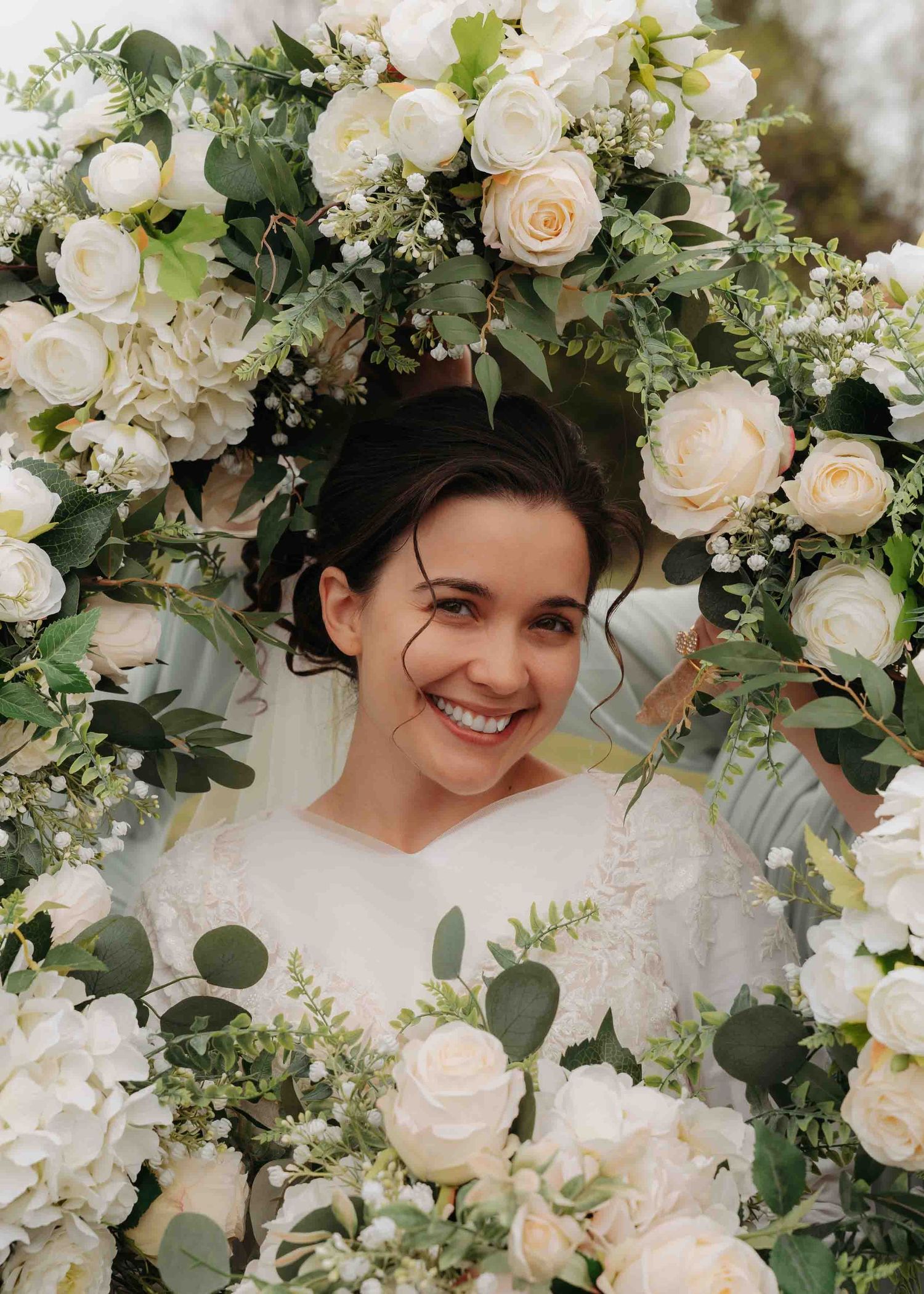 Smiling bride framed by a stunning arch of white roses and lush greenery on her wedding day.