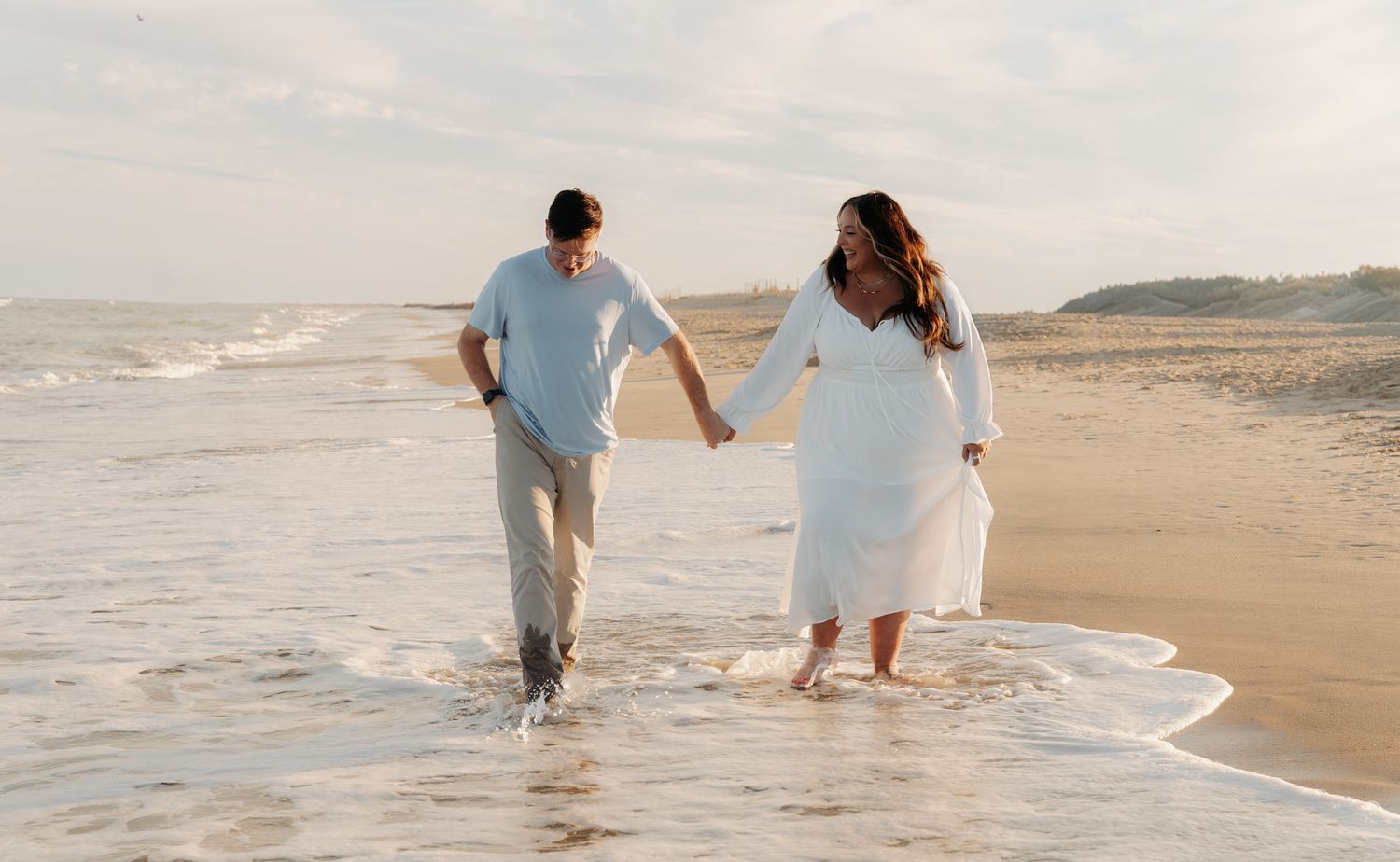 Couple holding hands walking along ocean shoreline, woman in white dress and man in casual attire at sunset.