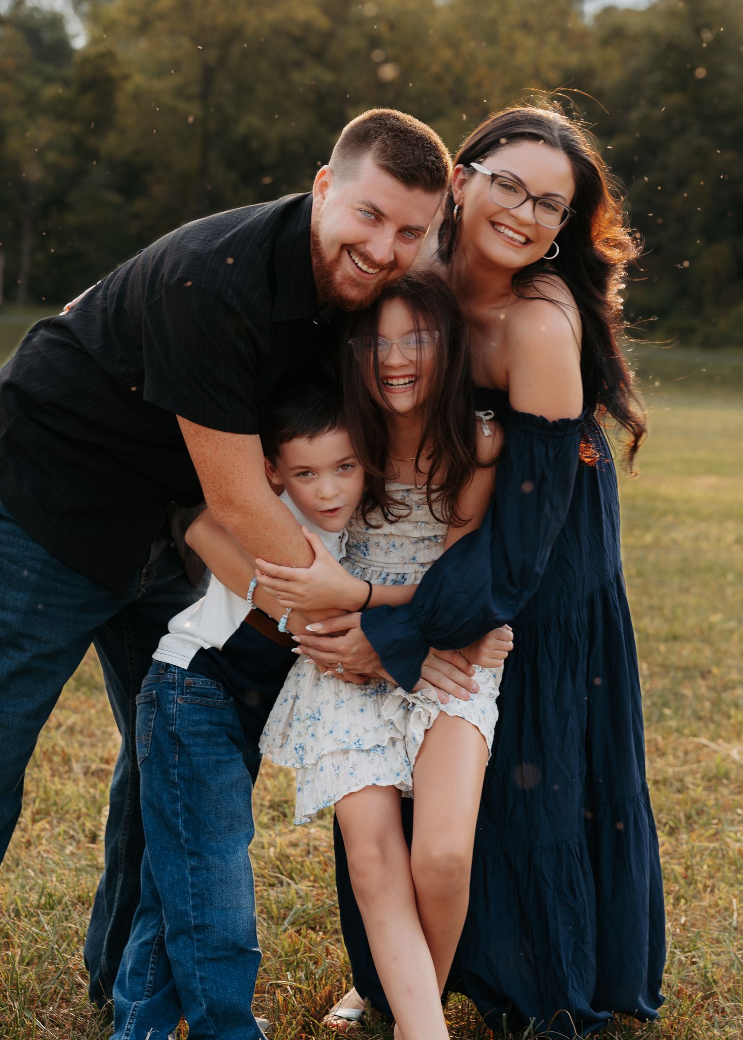 Laughing family of four hugging together in an outdoor field at golden hour.