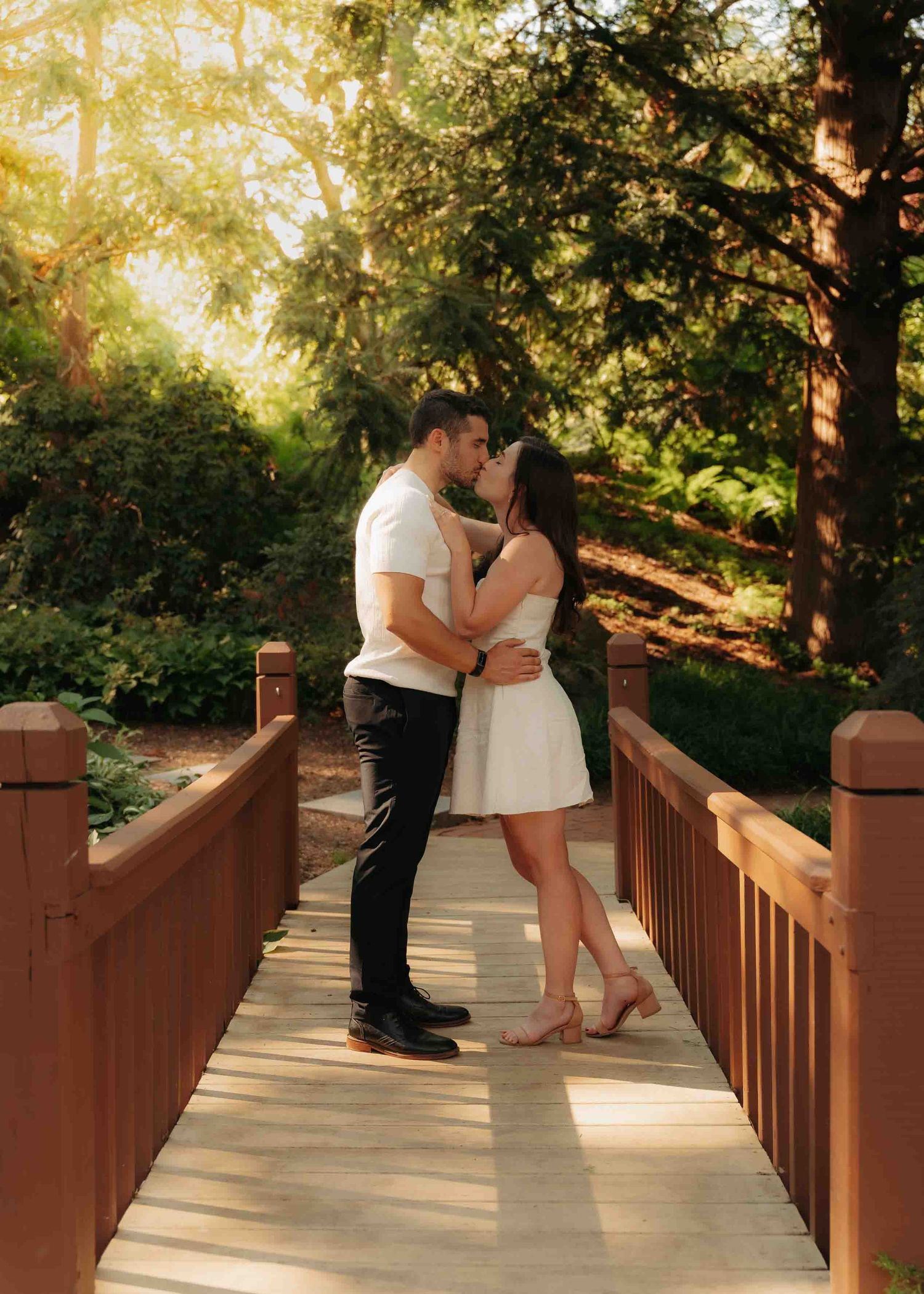 Couple sharing a kiss on a wooden bridge surrounded by lush green garden trees.