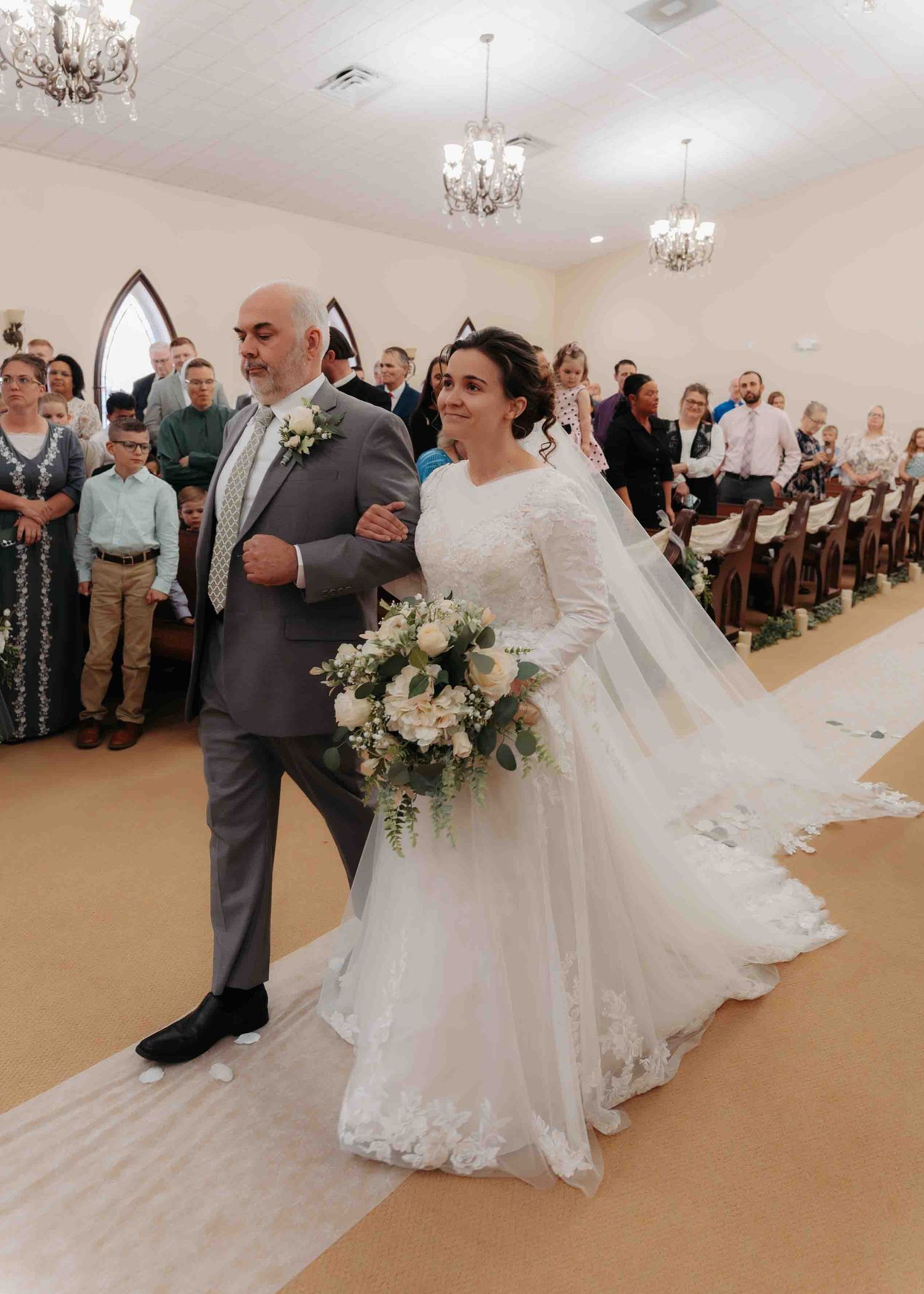 Father escorts his daughter in a long-sleeved bridal gown down the church aisle on wedding day.