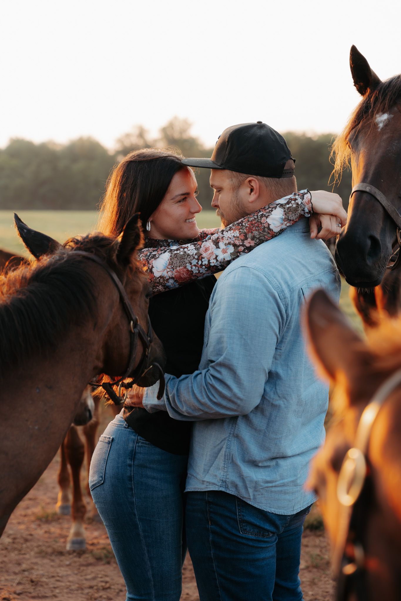Couple sharing a romantic moment surrounded by horses at golden hour on a scenic farm field.