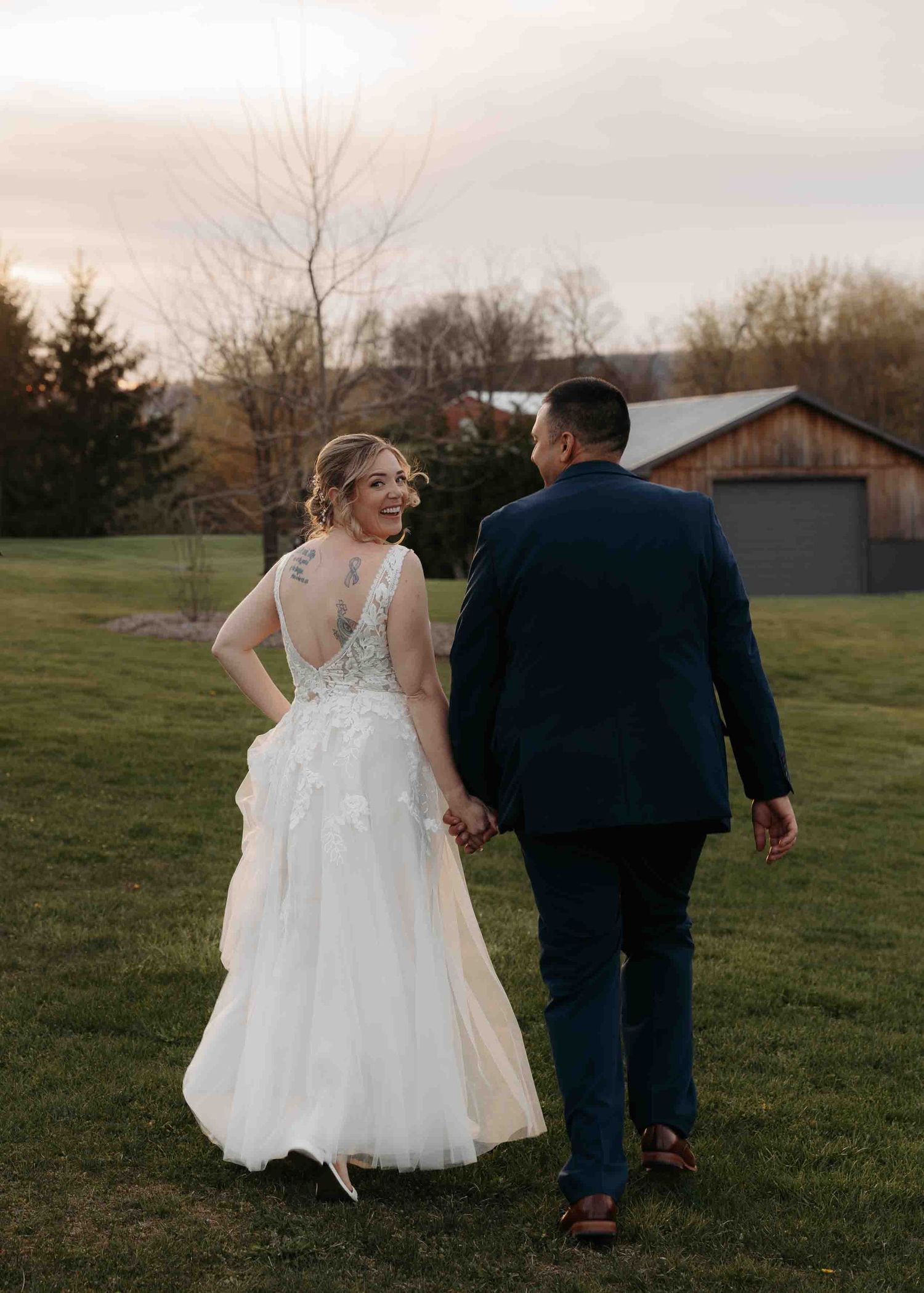 Bride in lace gown with back tattoo walks with groom at farm wedding sunset.