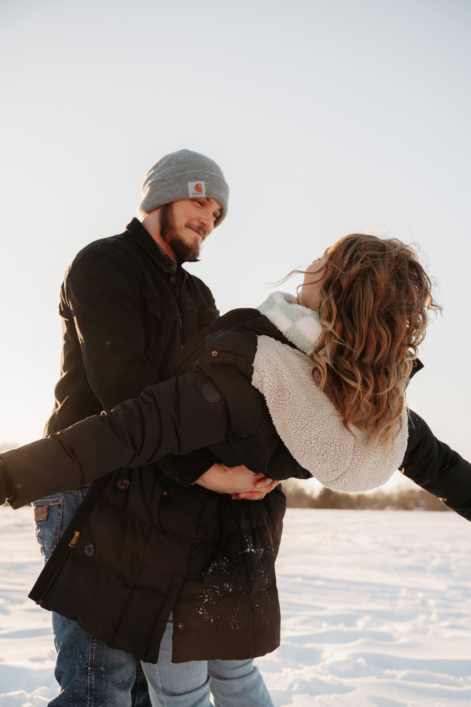 Man holding woman in his arms while smiling in a snowy winter landscape at sunset.