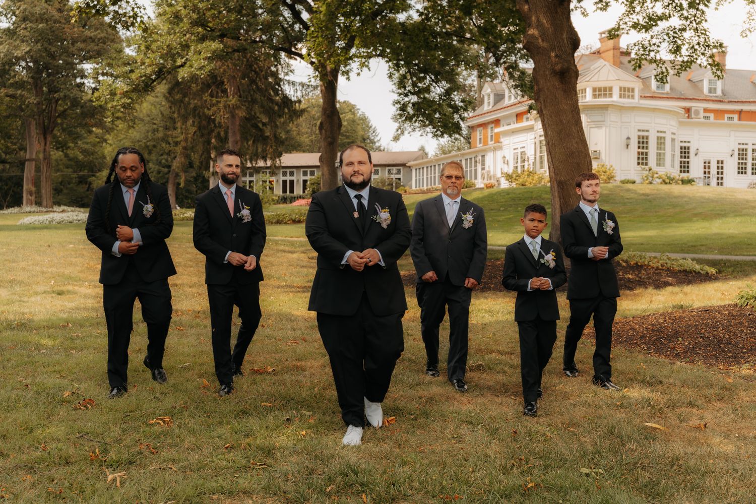 Groom and groomsmen in black suits walking confidently across a green lawn at wedding.