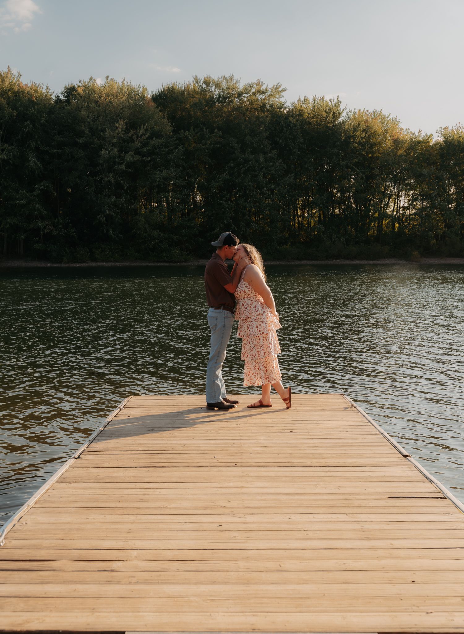 Couple sharing a romantic kiss on a wooden dock overlooking a calm lake surrounded by trees.