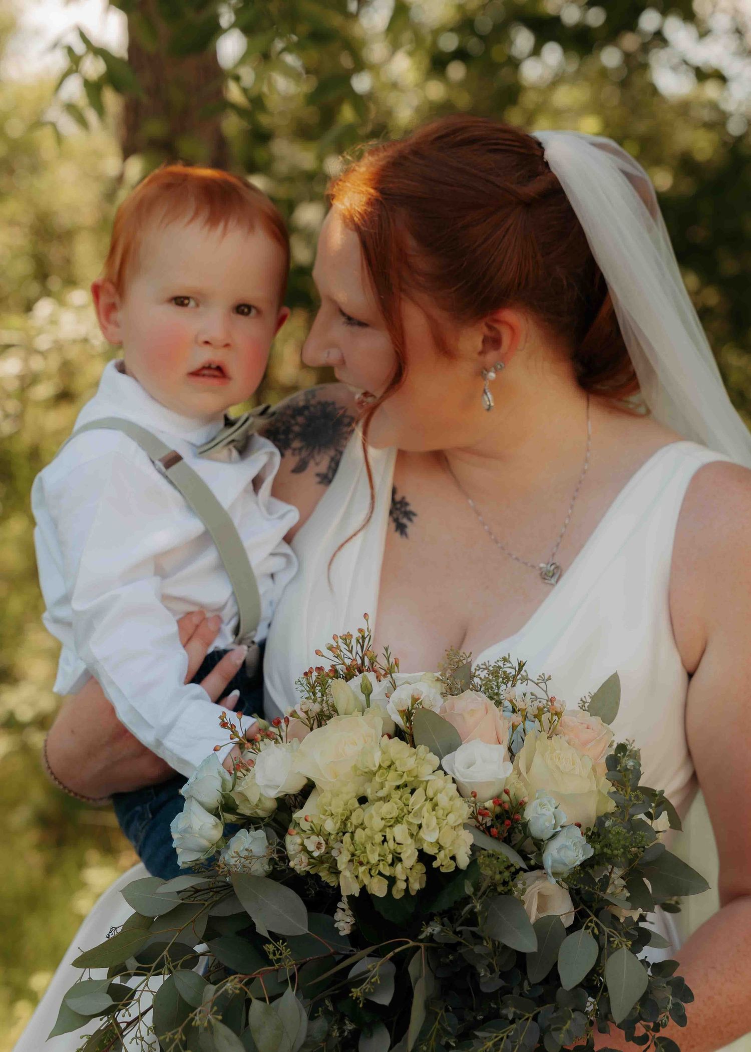 Bride in white gown holding bouquet of white roses and greenery while kissing a red-haired baby boy in suspenders.