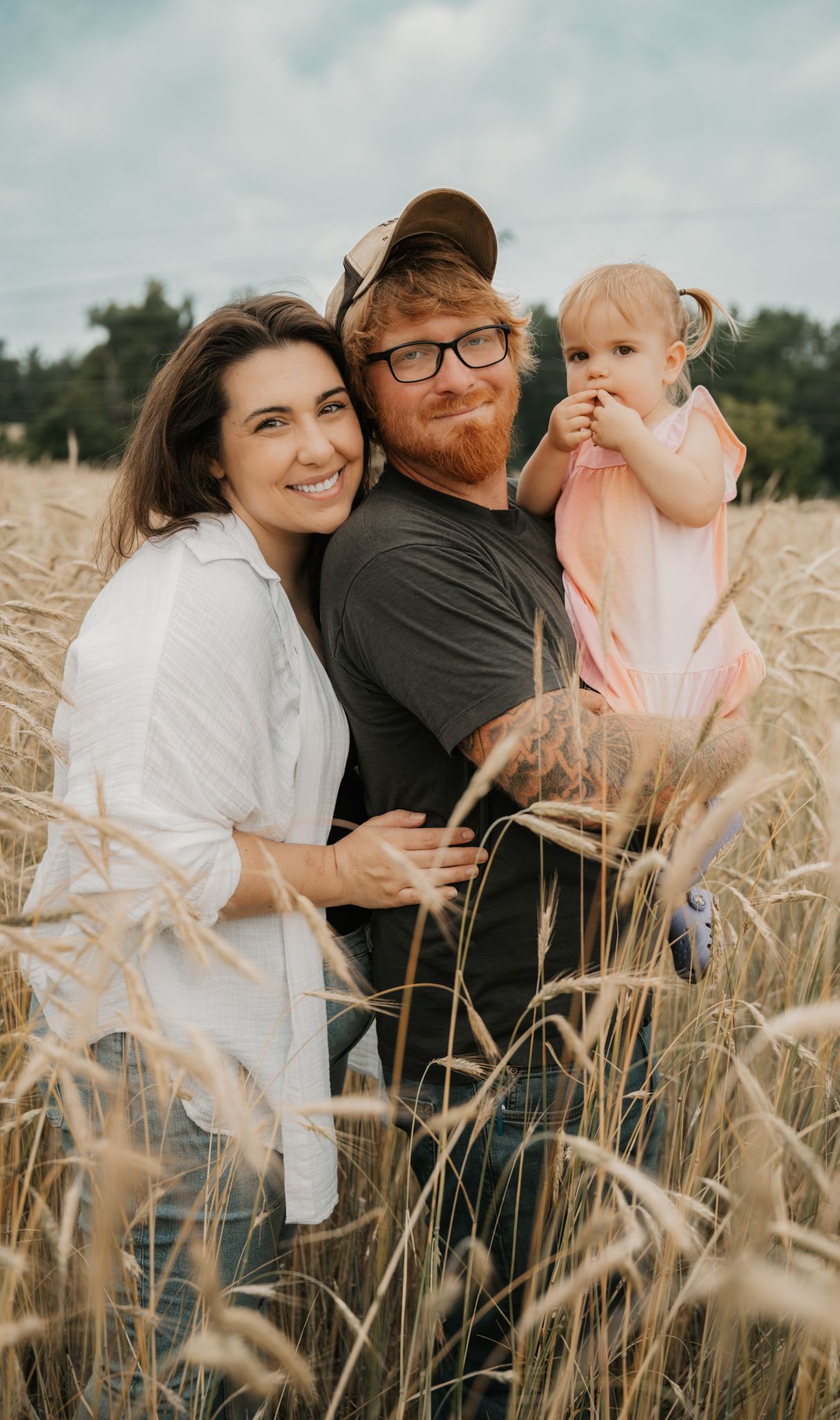 Happy family of three posing in a golden wheat field, parents smiling with baby girl.