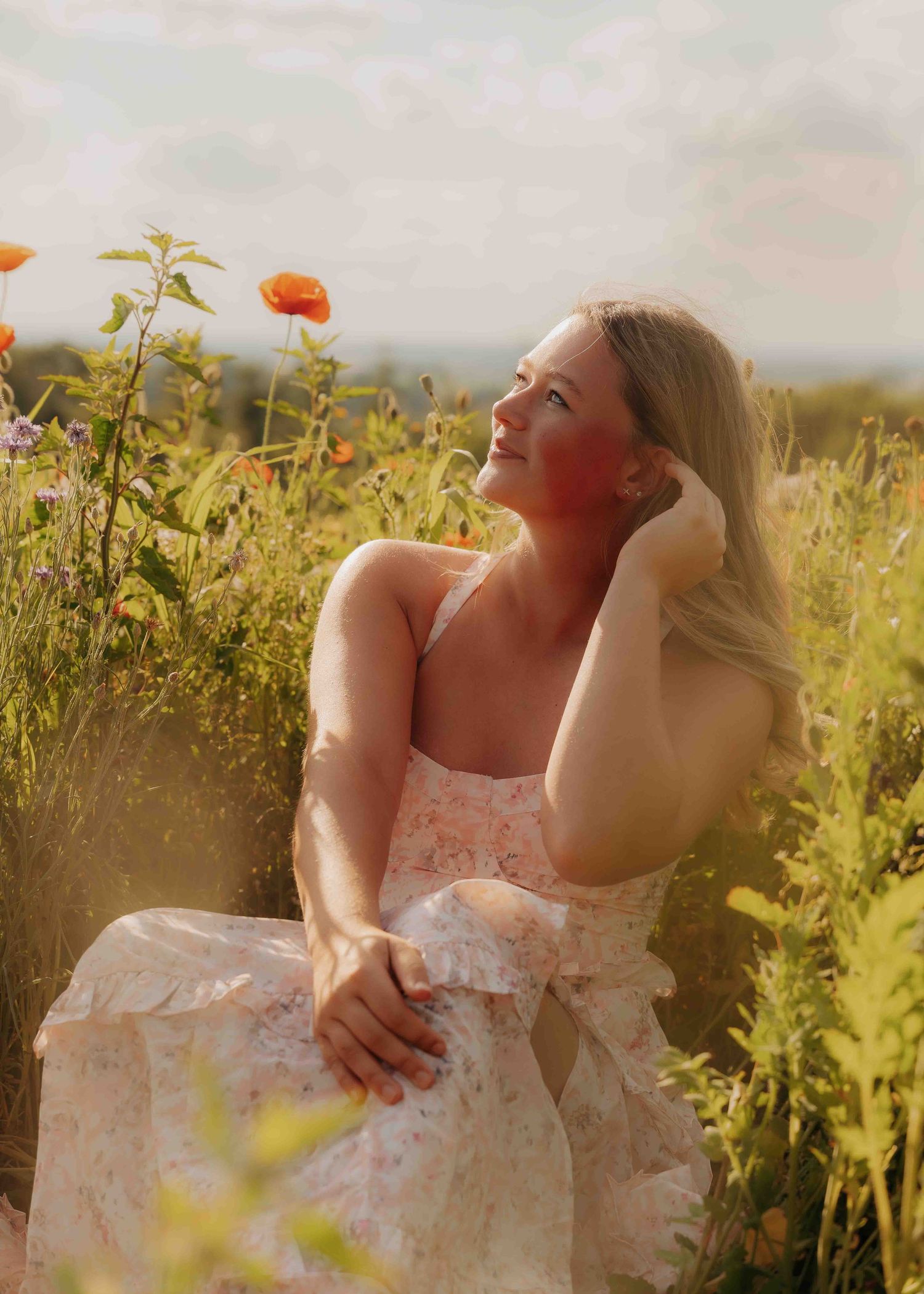 Young blonde woman in floral dress sitting in a sunlit poppy field, smiling up at the sky.