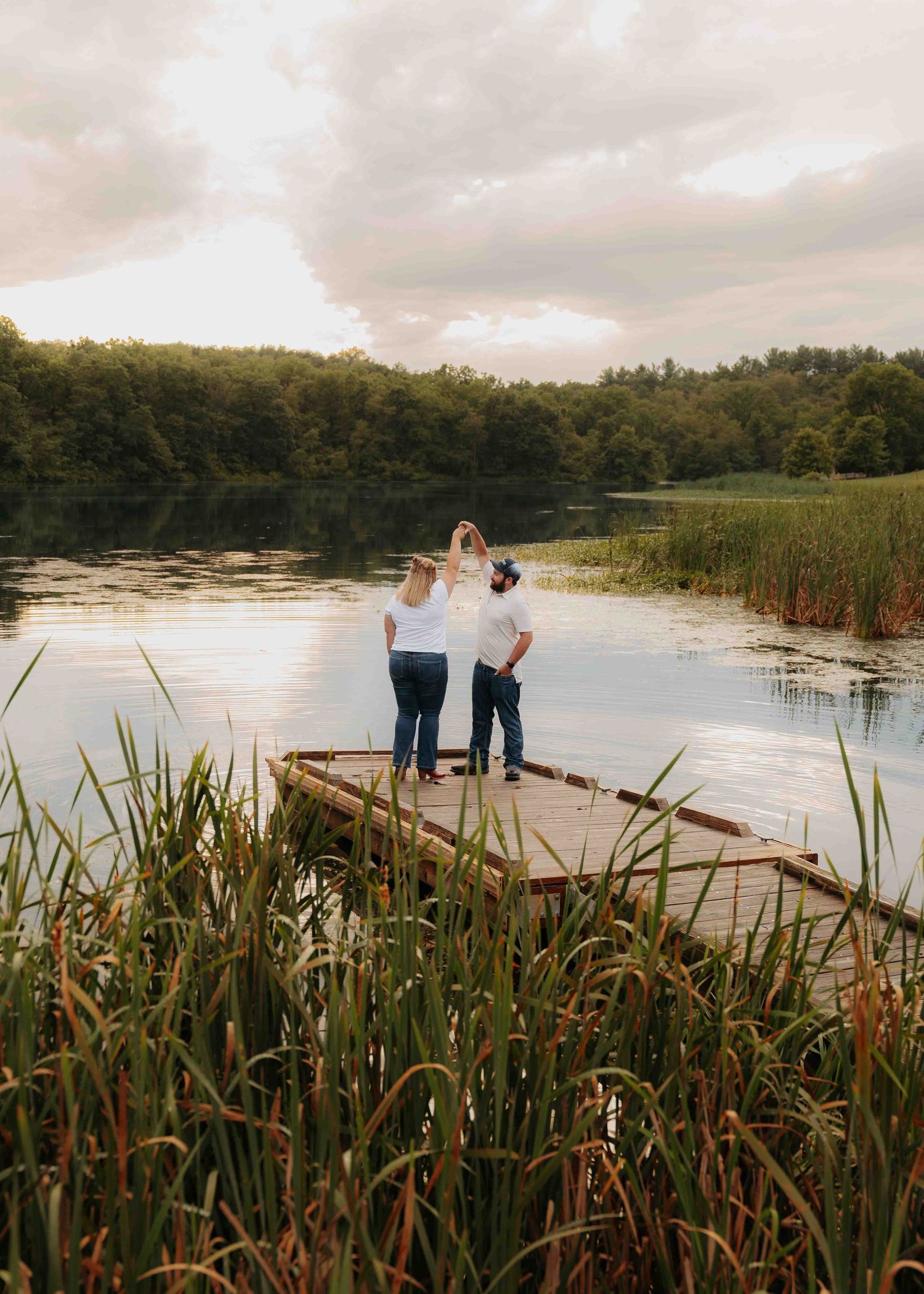 Three people stand on a wooden dock surrounded by reeds on a calm lake at sunset with lush green trees in the background.