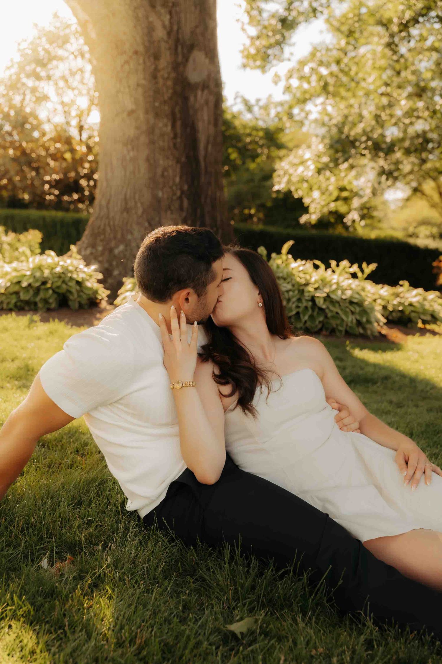 Couple sharing a kiss while sitting on grass beneath a large tree in a sunlit garden.