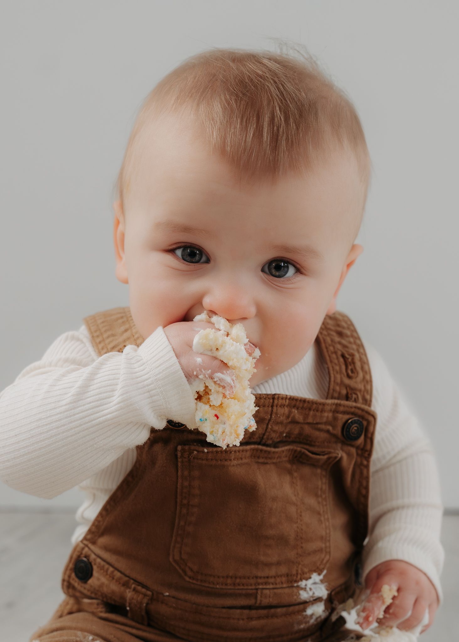 Adorable baby in brown overalls eating cake during a first birthday smash session.