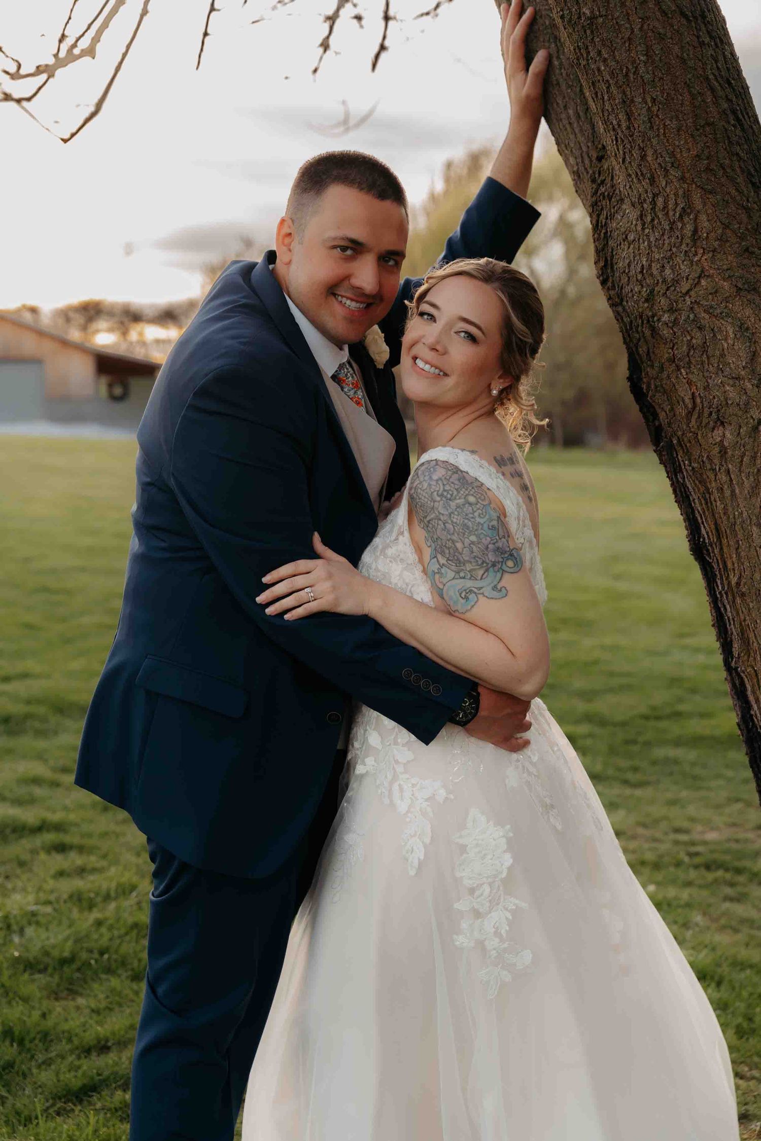 Happy bride and groom embracing by tree at outdoor farm wedding at sunset.