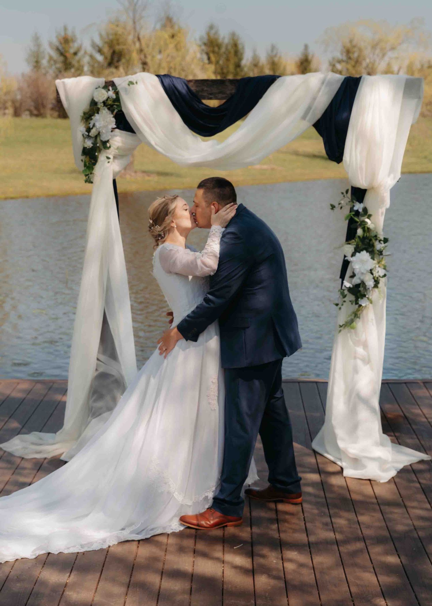 Bride and groom share first kiss under floral wedding arch draped with navy and white fabric by a serene lake.