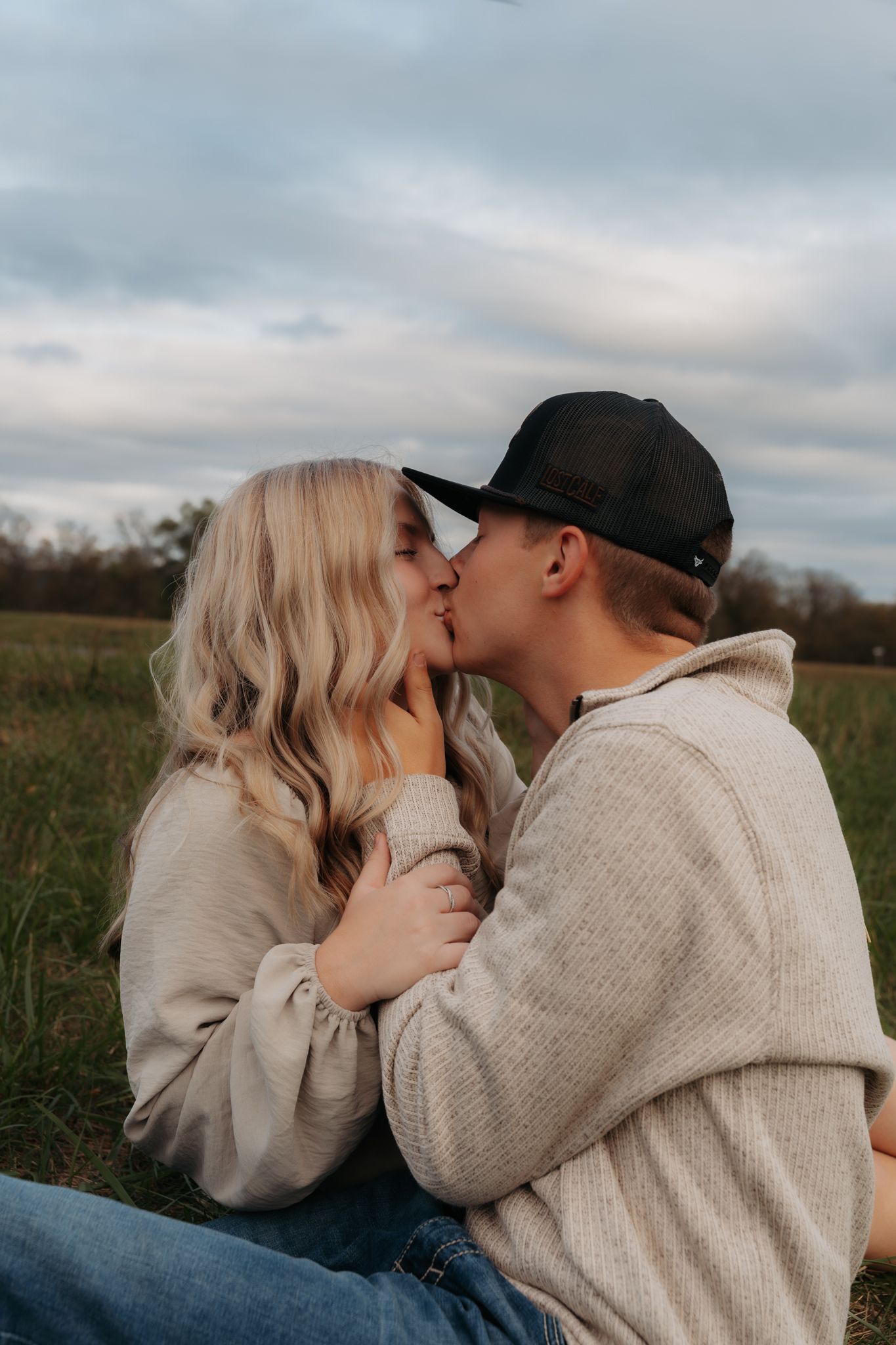 Young couple sharing a kiss while sitting in a green field under a cloudy sky.