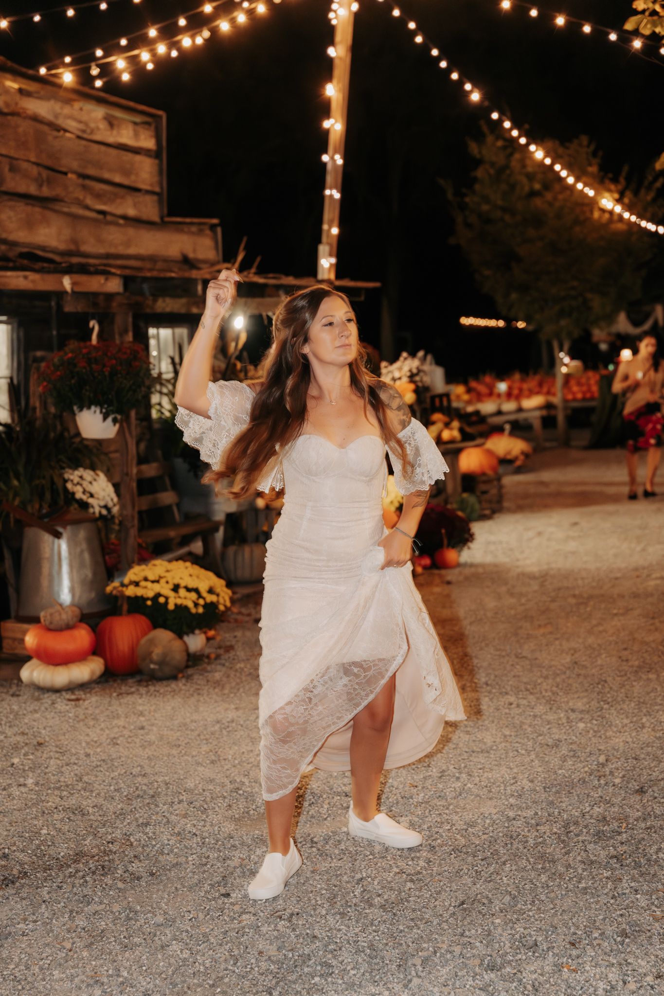 A bride in a white lace dress twirls joyfully under string lights at a rustic outdoor fall wedding venue at night.
