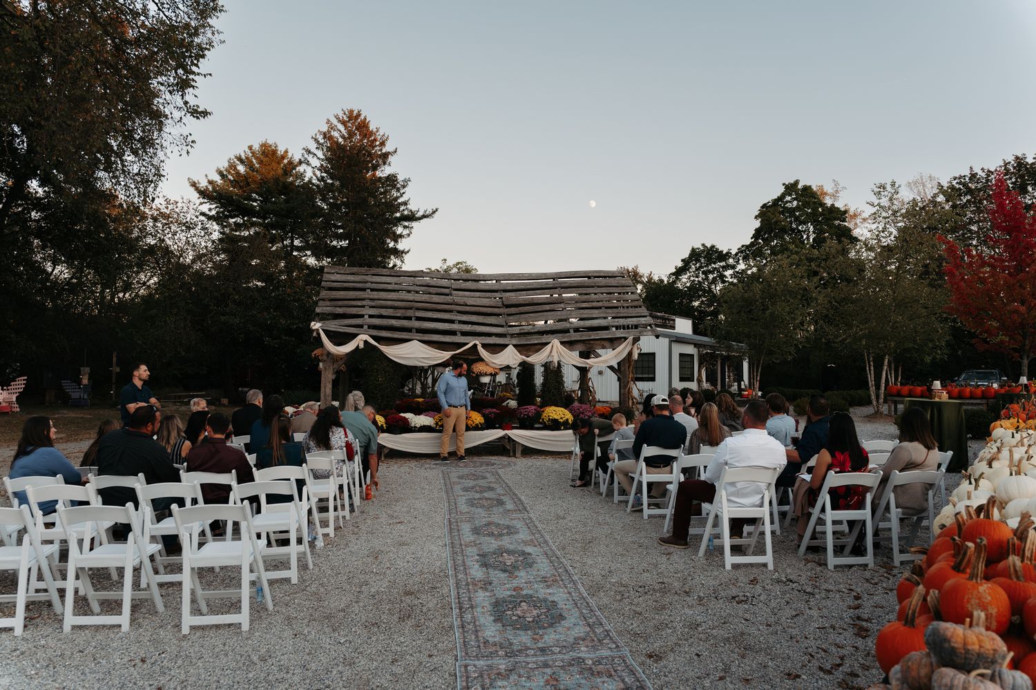 Outdoor fall wedding ceremony at dusk with white chairs and pumpkin decorations.