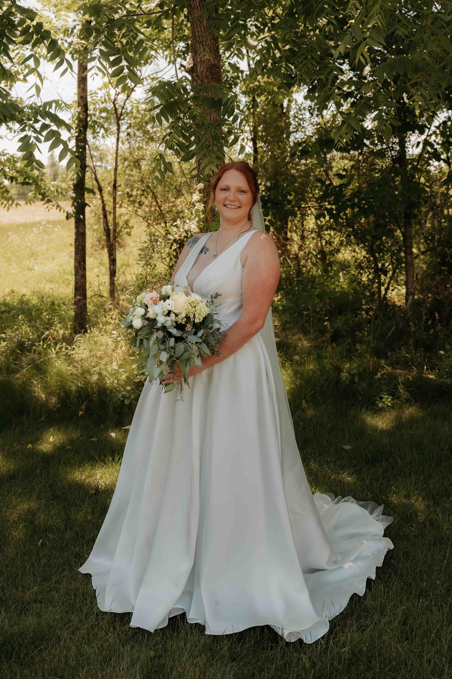 Bride in elegant white A-line gown holding bouquet of white flowers in lush green outdoor setting.