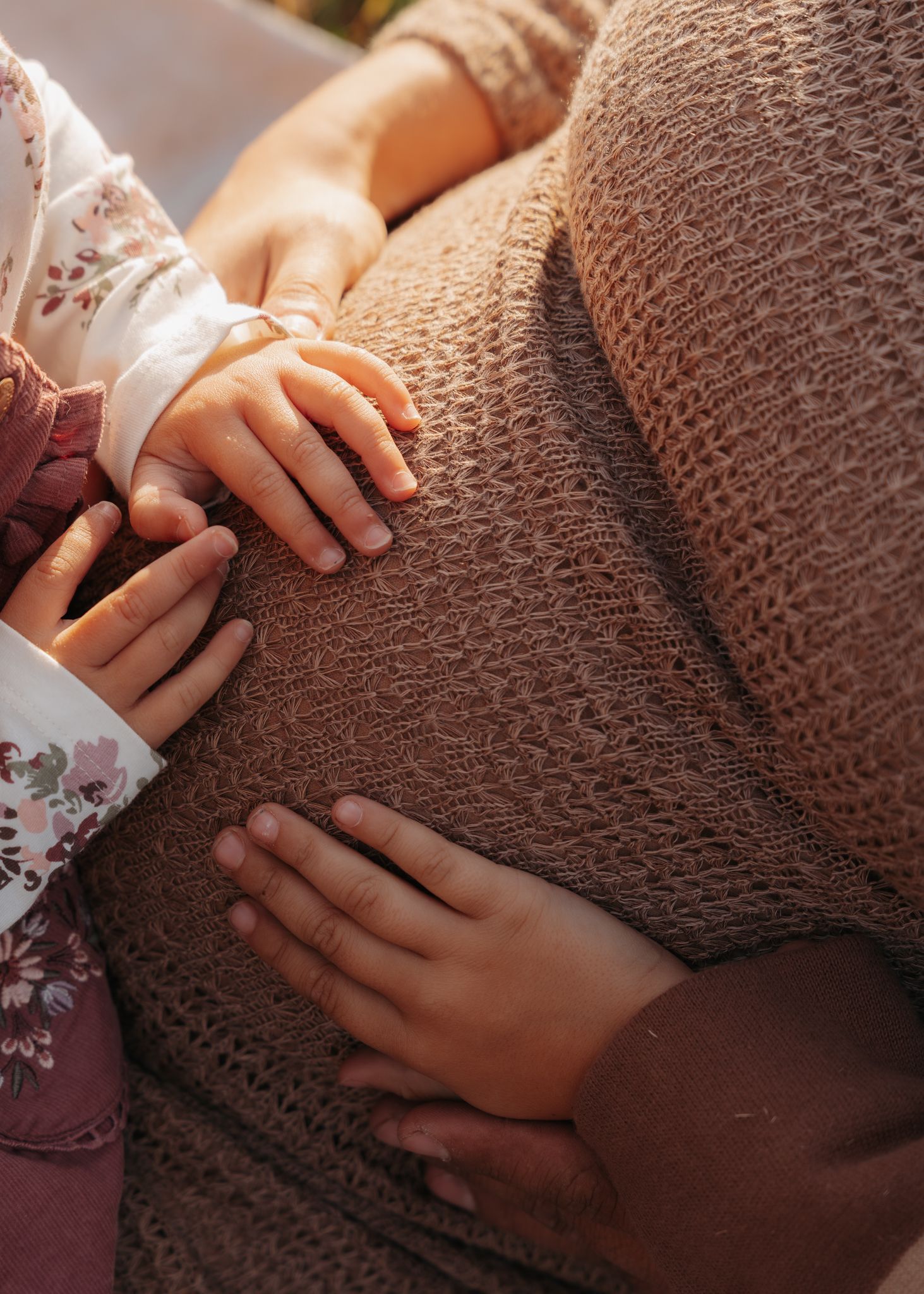 Close-up of child and adult hands resting on pregnant mothers belly outdoors.