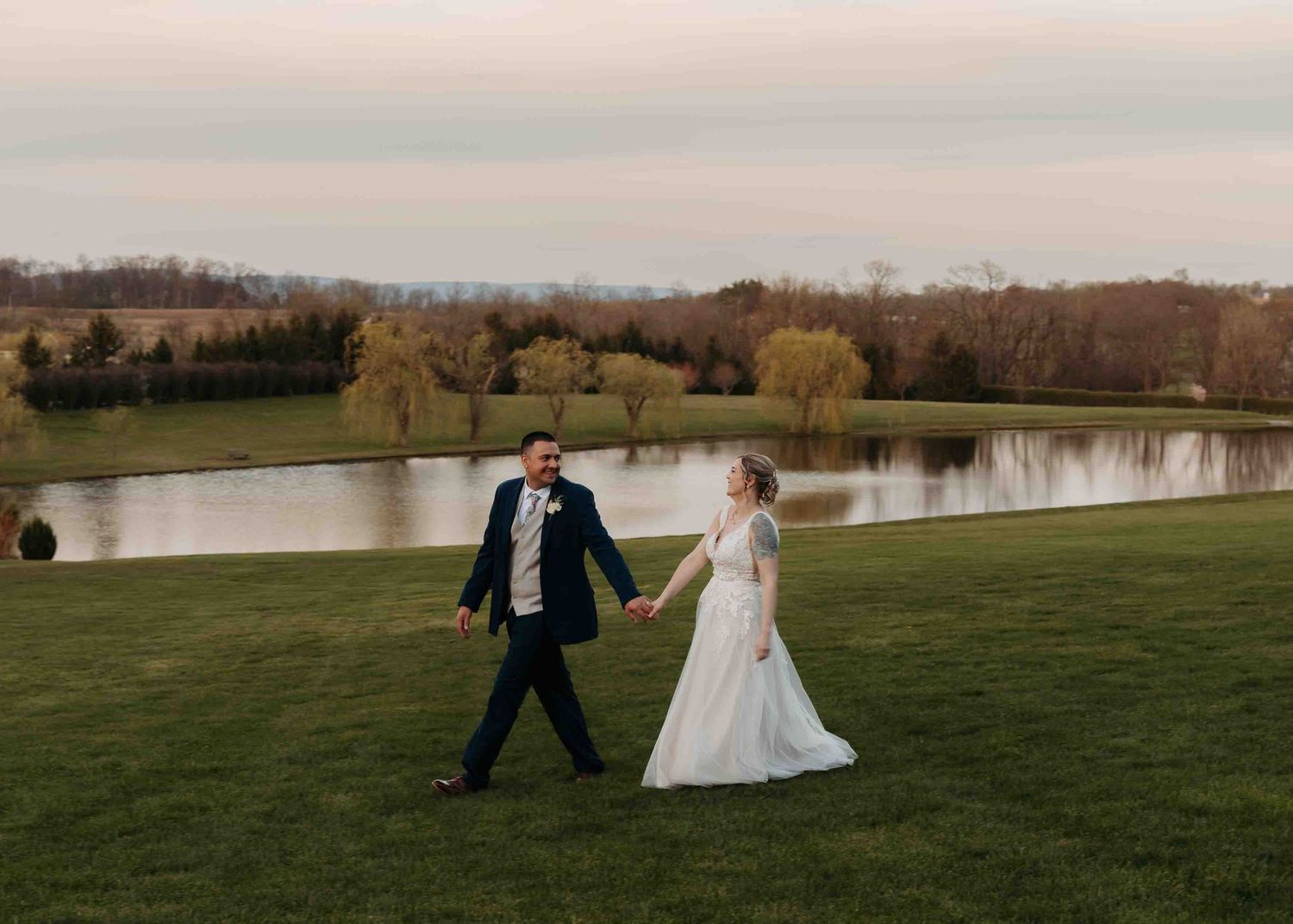 Bride and groom holding hands walking near scenic pond at outdoor farm wedding.